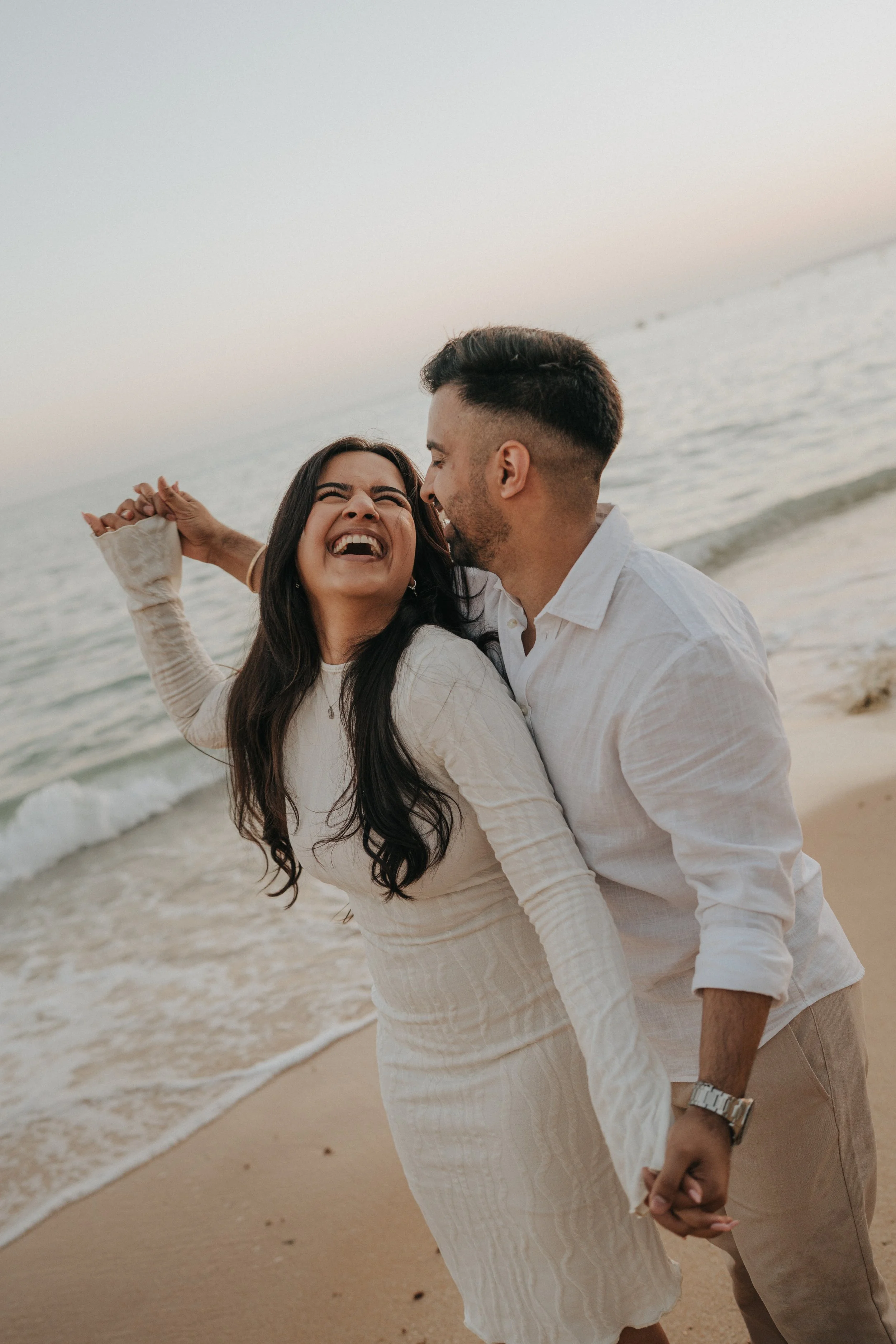 A smiling woman and a man holding hands and dancing on the beach during sunset, near the ocean waves.