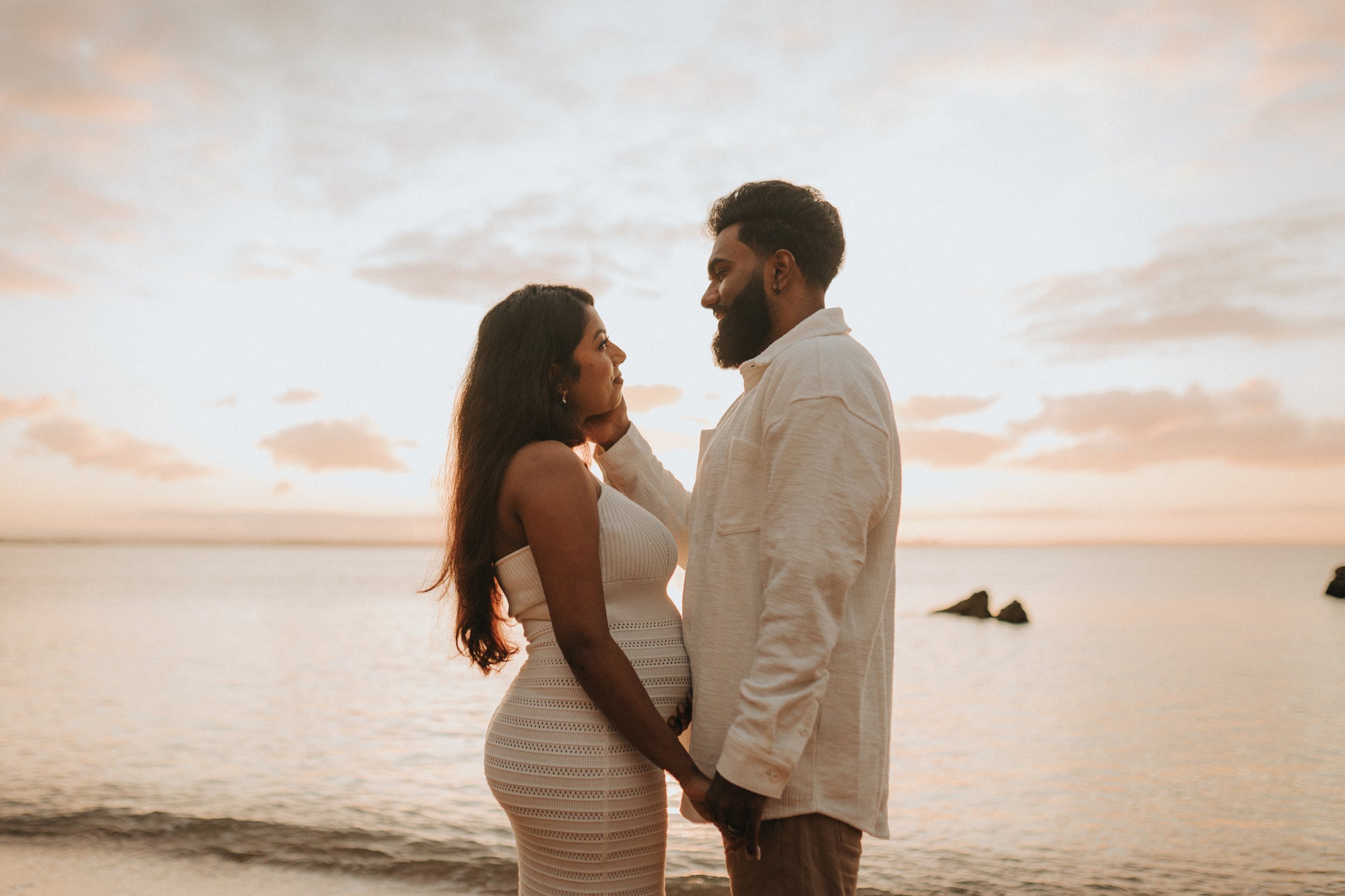 A happy couple stands close together on a beach at sunset, gazing into each other's eyes and holding hands. The woman has long dark hair and wears a white, sleeveless, fitted dress. The man has a beard and short hair, dressed in a light-colored shirt