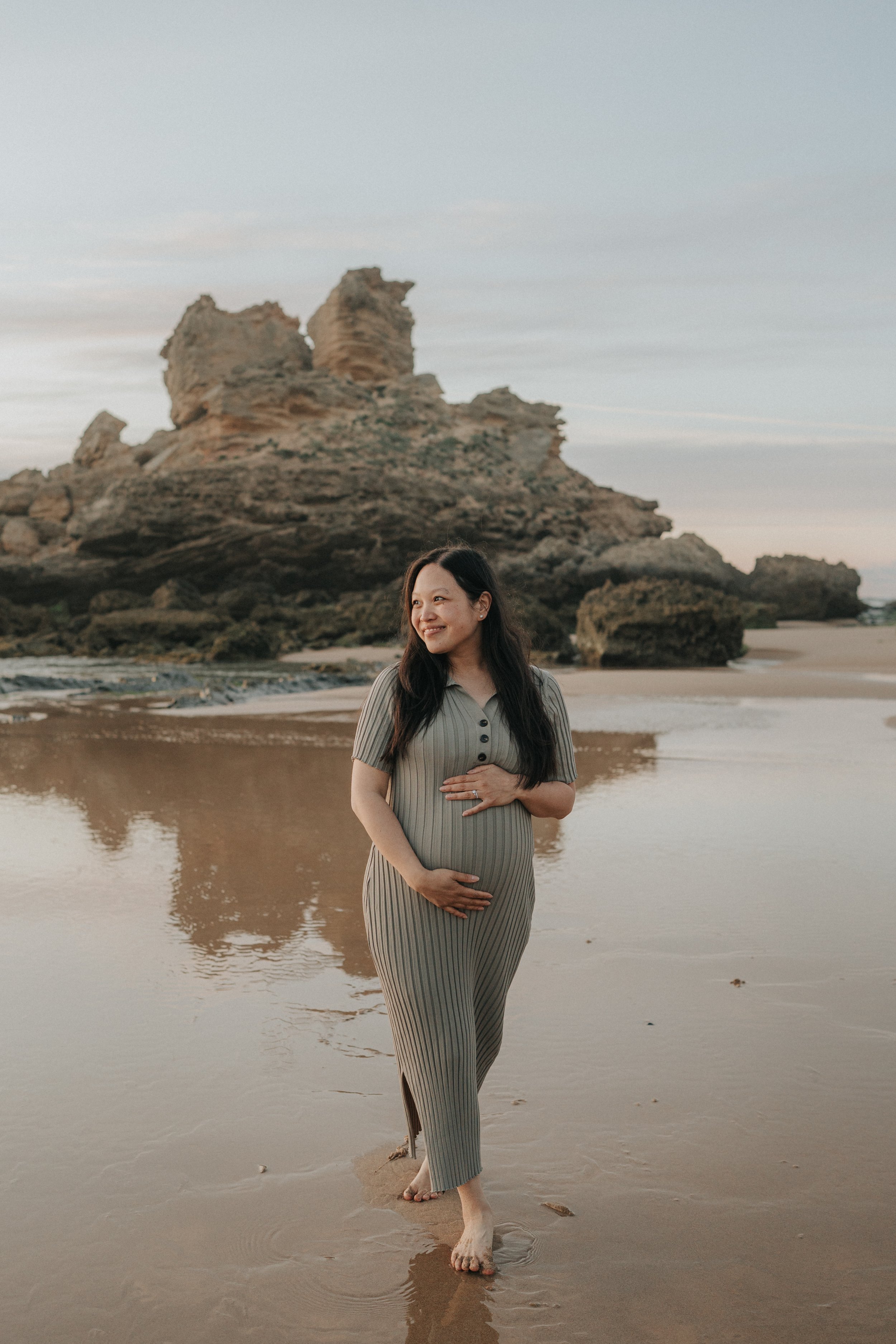 A pregnant woman in a beige ribbed dress standing barefoot on a sandy beach, holding her belly with her hands, smiling, with large rock formations in the background and calm water reflecting her image.