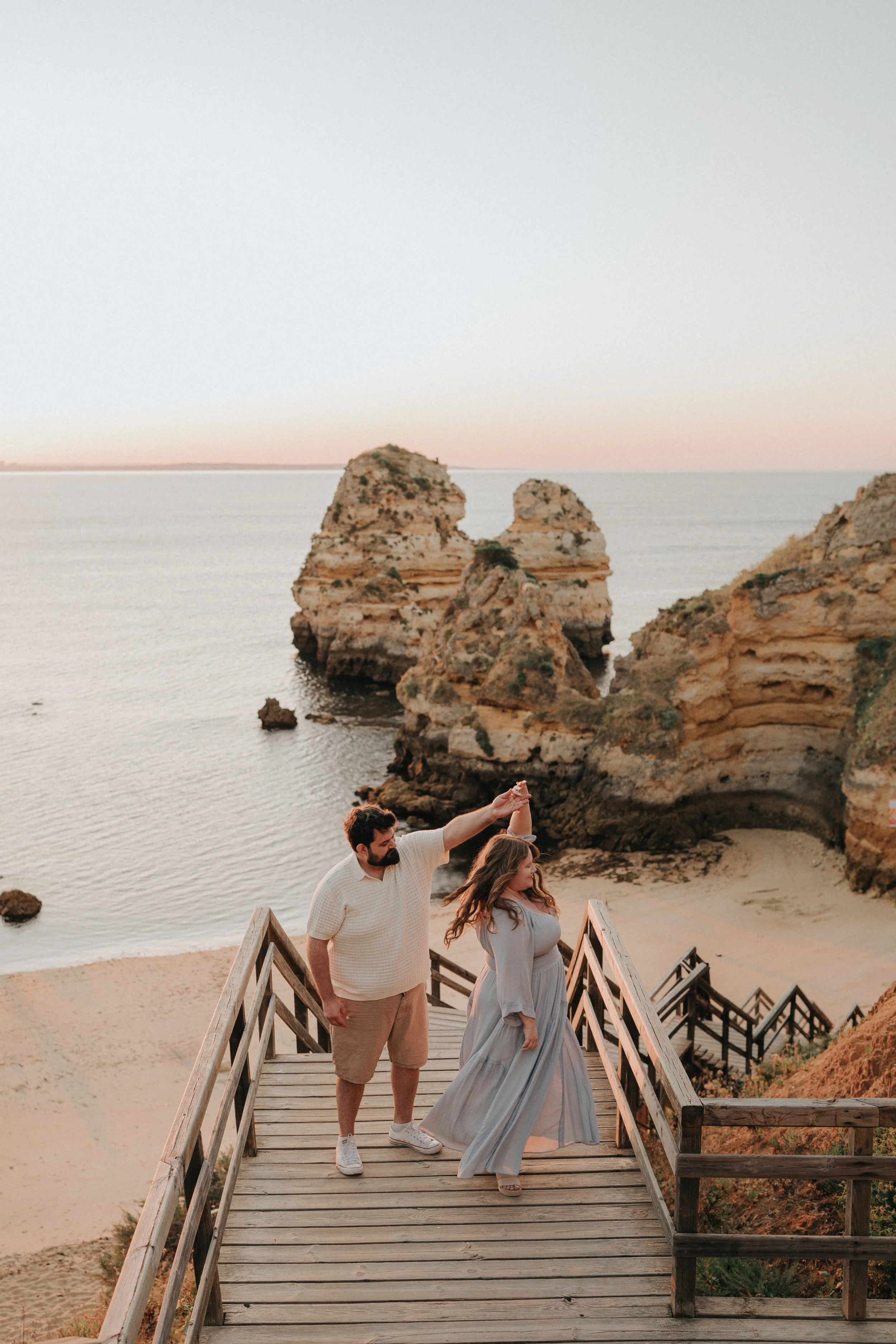 A couple dancing on a wooden staircase leading down to a beach with rocks and water, with large rocks in the background under a pastel sky.