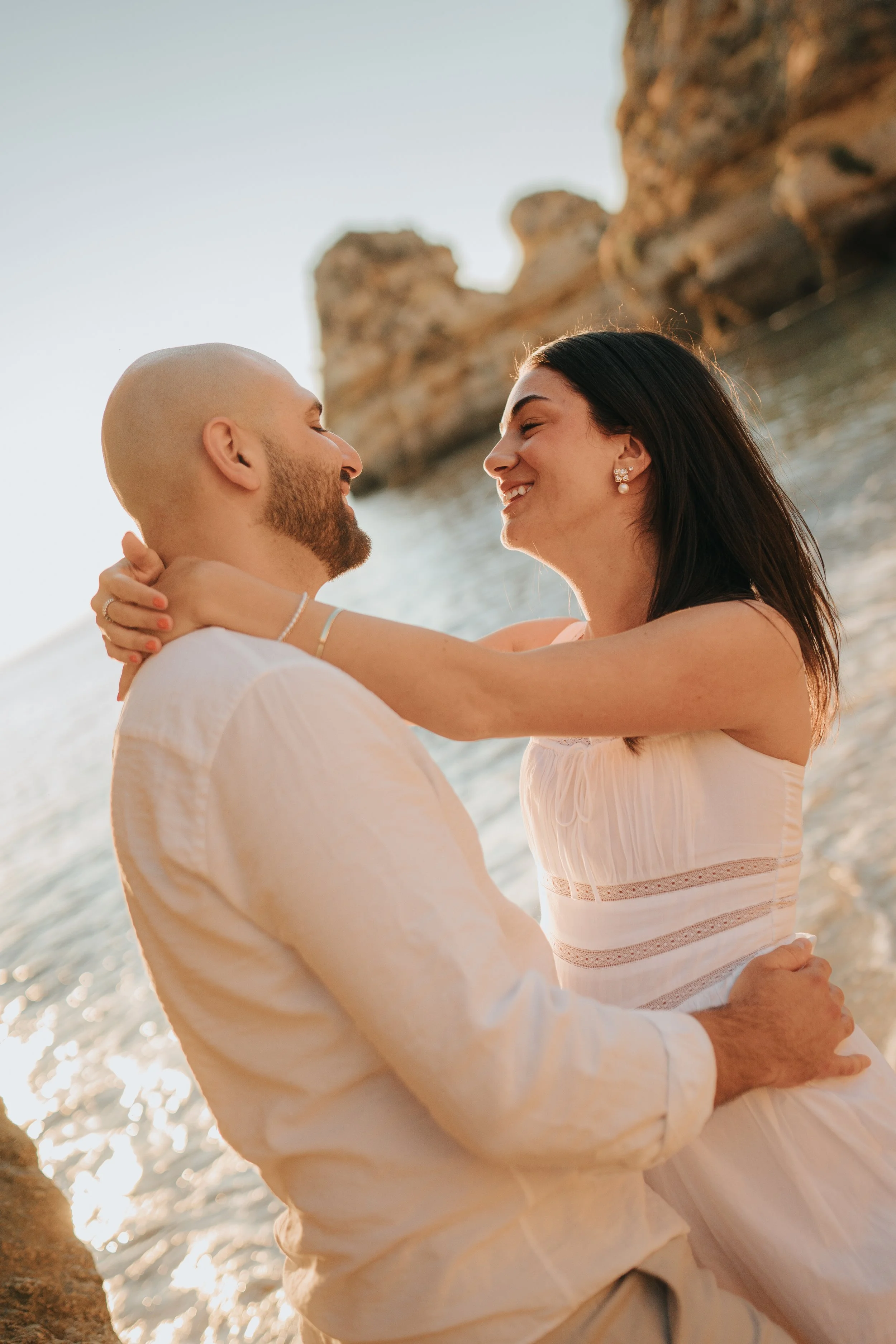 A couple embracing on a beach with rocky cliffs in the background, smiling at each other during sunset.