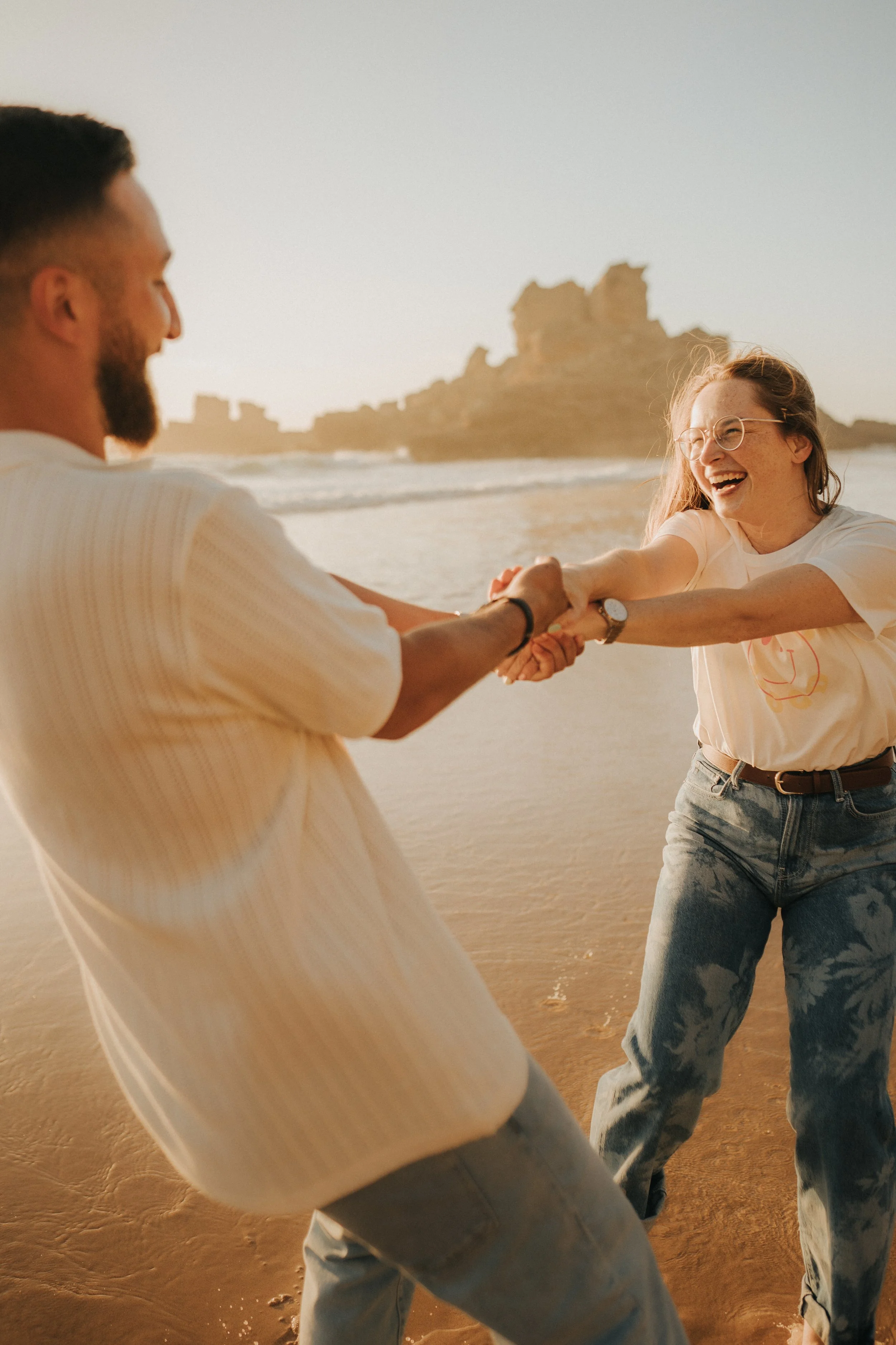 Two people holding hands and laughing on the beach during sunset with rocky formations in the background.