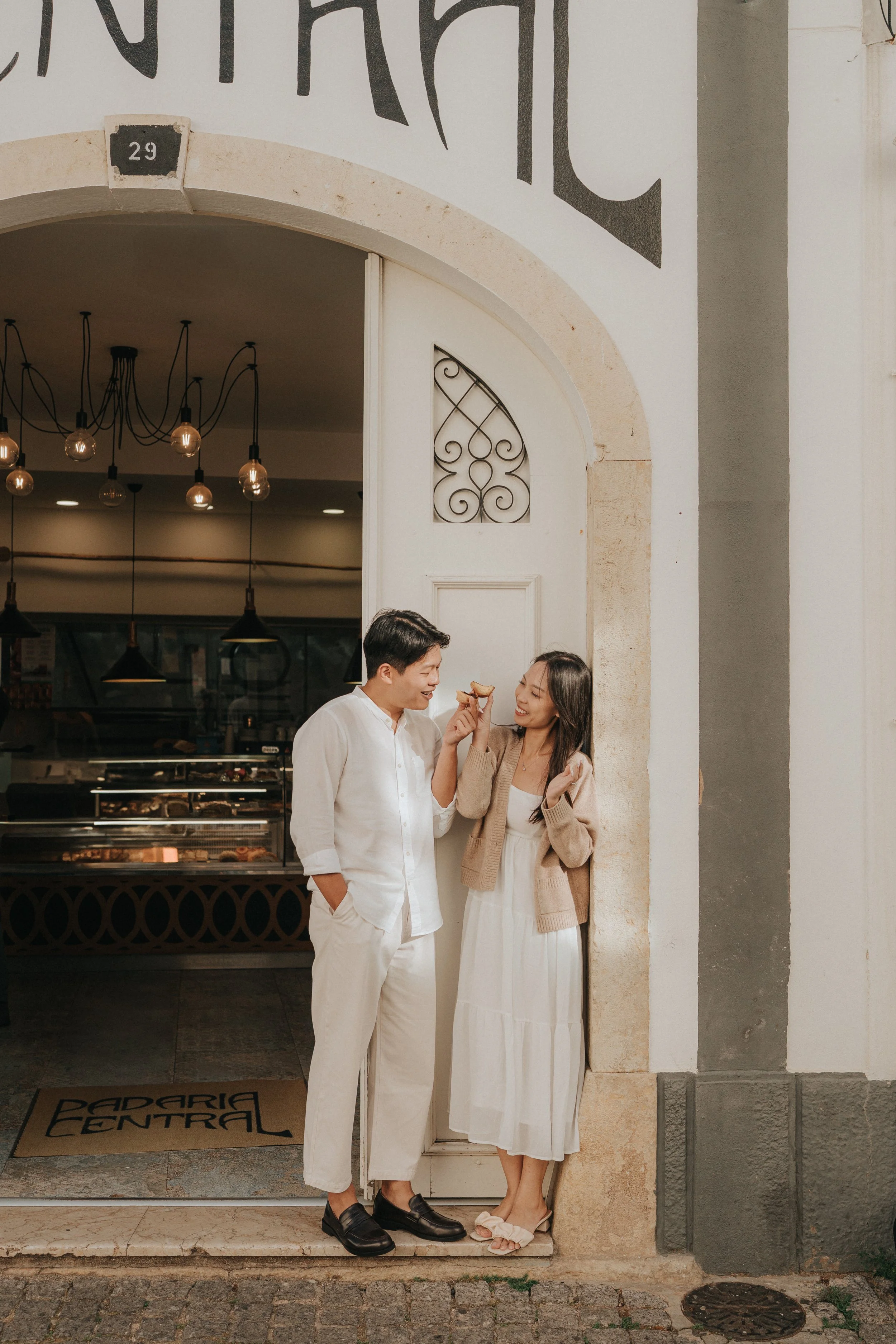 A man and woman standing at the entrance of a restaurant, smiling and sharing a piece of food. The man is dressed in a white shirt and beige pants, while the woman wears a beige cardigan and a white dress. The restaurant sign above them reads '29' an
