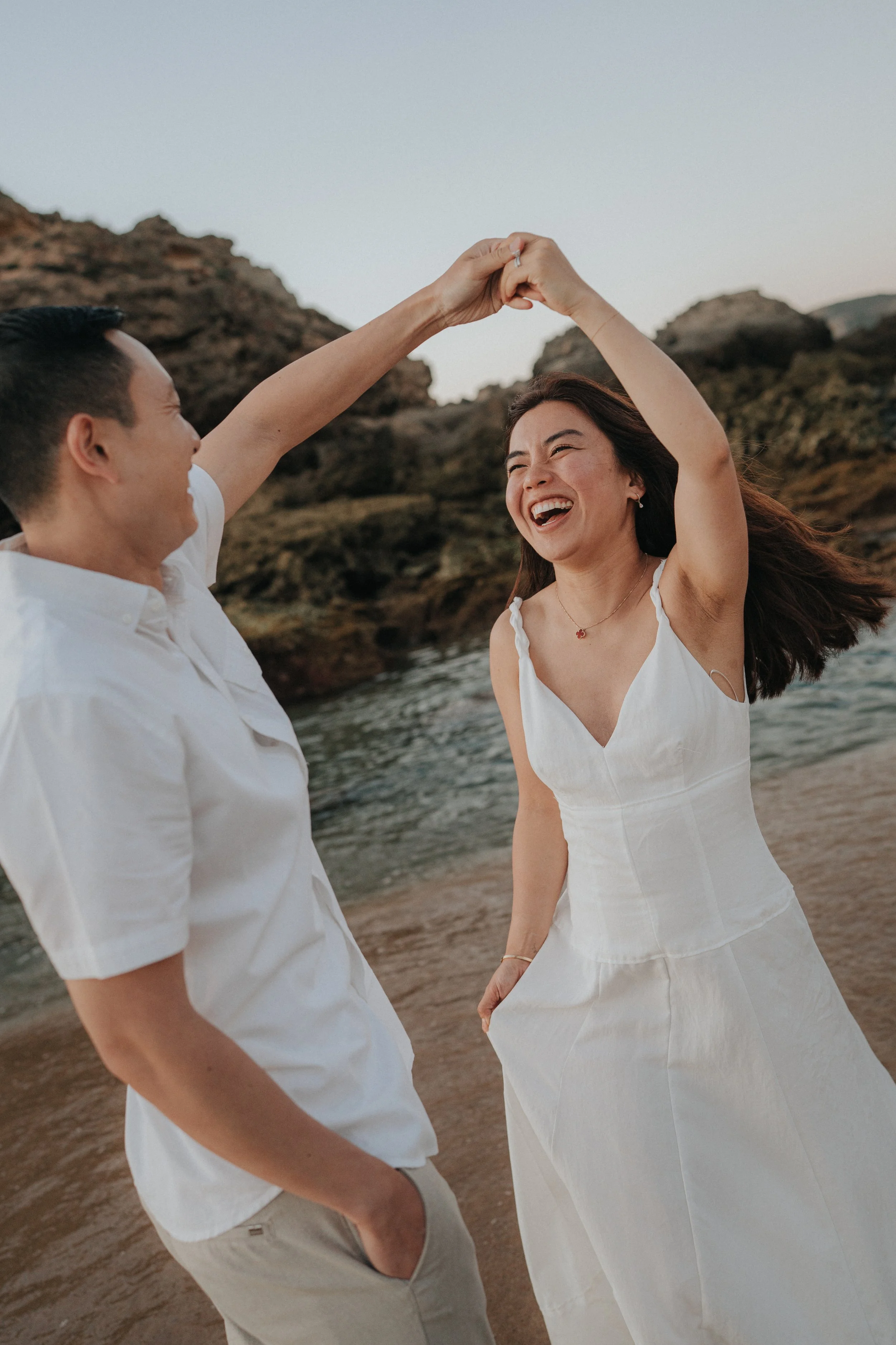 A man and woman holding hands and twirling on a beach, smiling happily, with rocky formations and the ocean in the background during sunset.