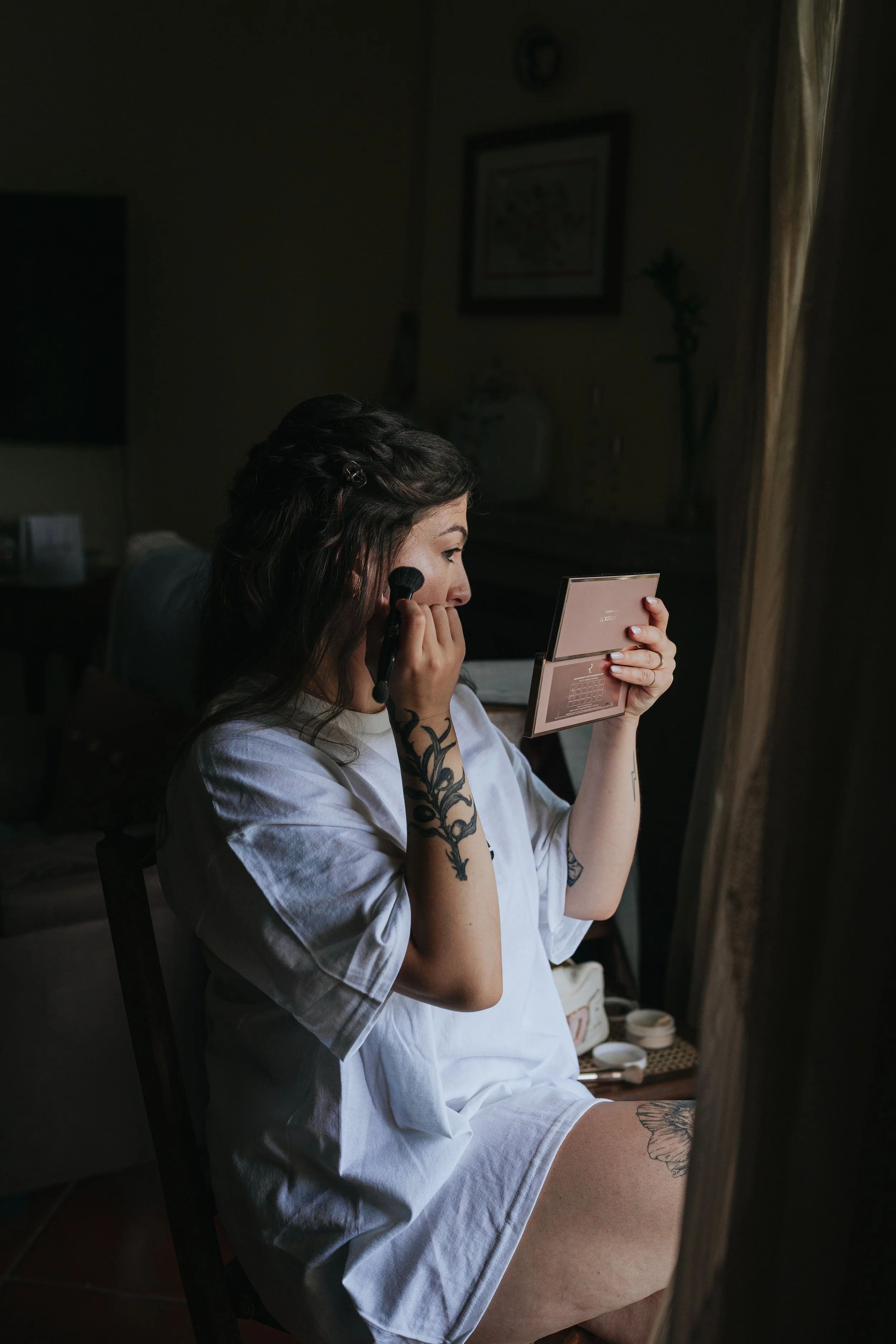 A woman with tattoos on her left arm and thigh applying makeup while looking into a pink compact mirror in a dimly lit room.