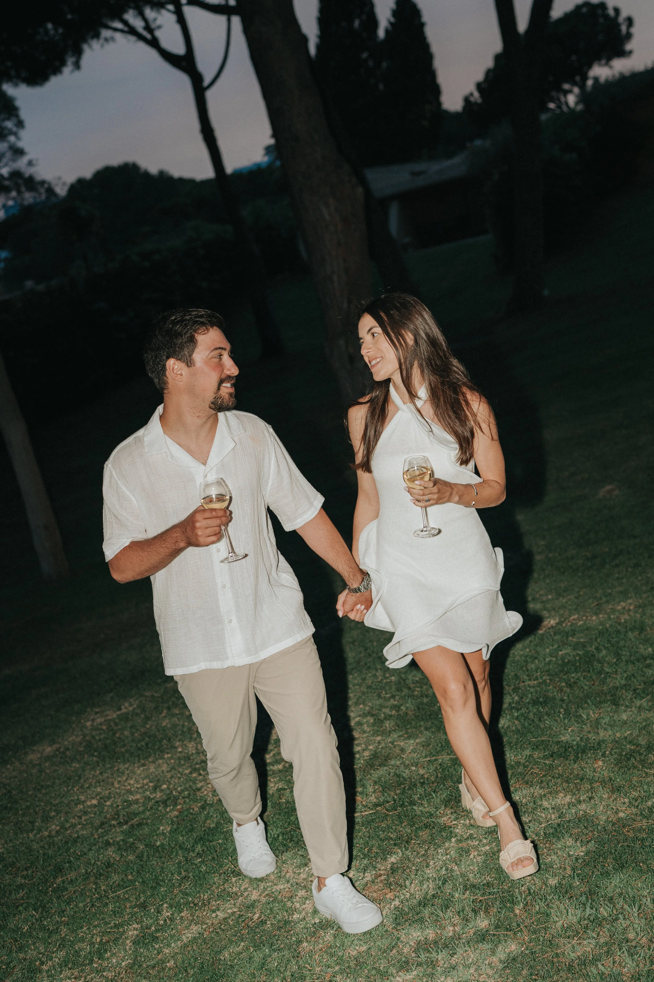 A man and woman walk hand in hand on a grassy area during dusk, both holding glasses of white wine, smiling at each other.