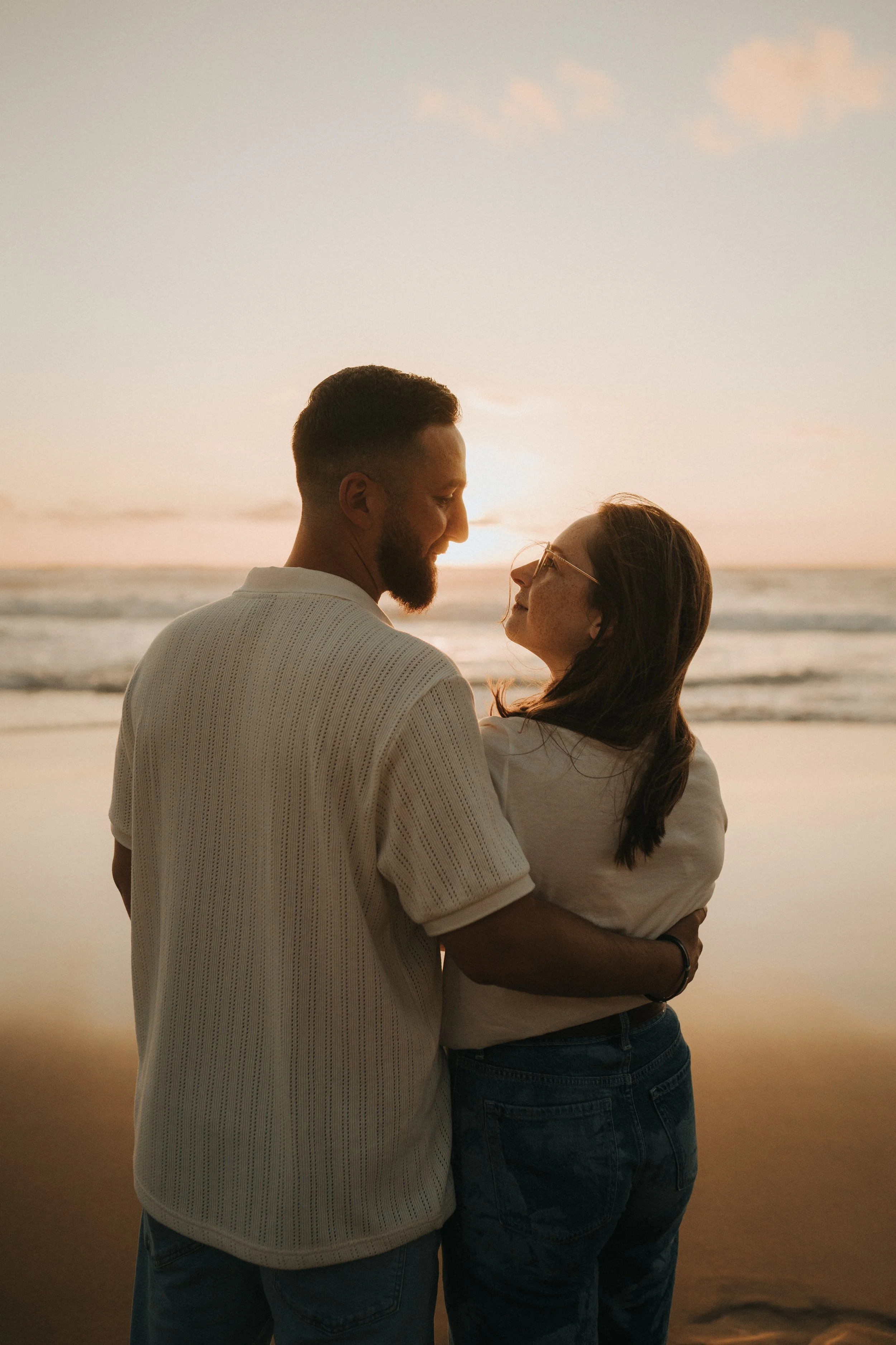 A man and woman standing close together on a beach at sunset, facing each other and smiling.