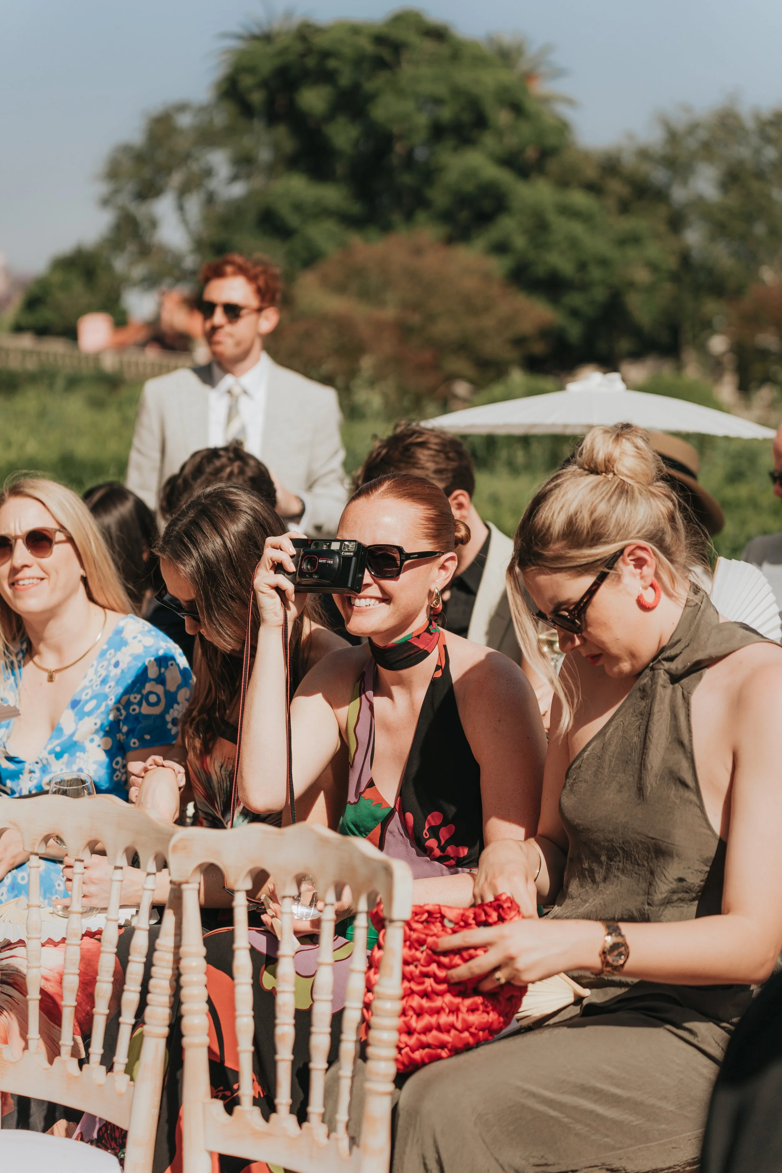 People gathering outdoors at a daytime event, some wearing sunglasses and colorful clothing, with one person taking a photo with a camera, others seated, and a man in a light-colored suit and sunglasses standing in the background.