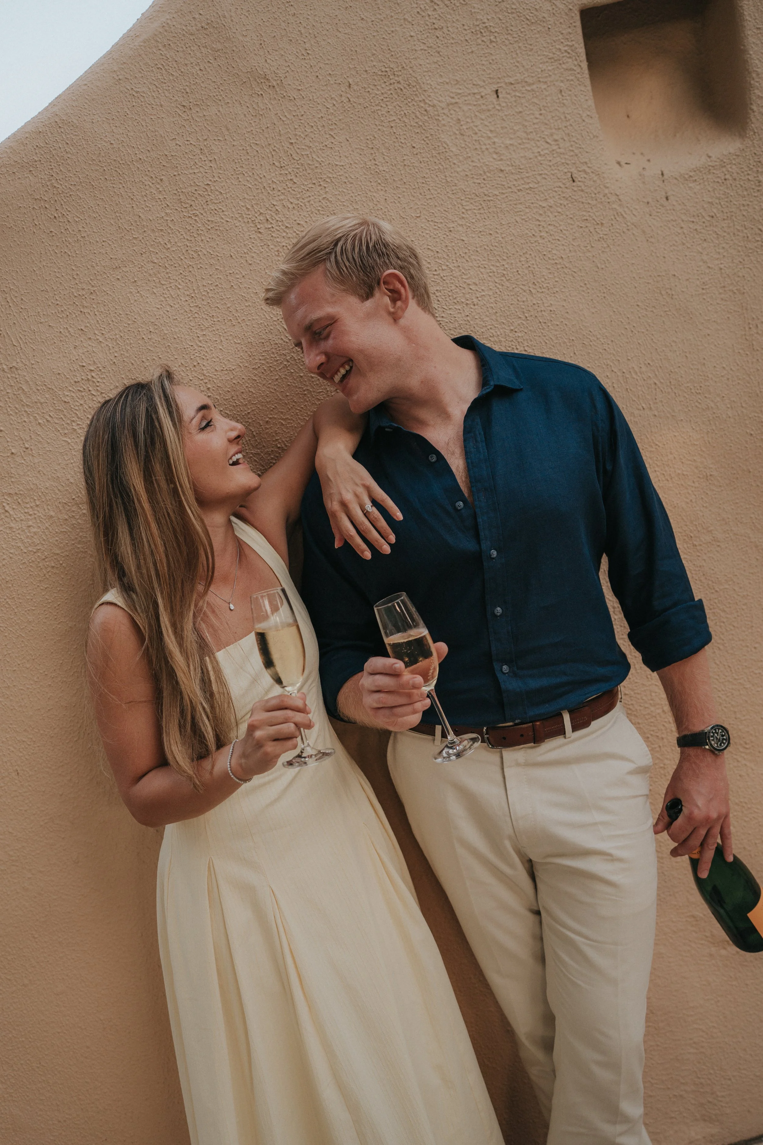 A man and woman smiling and looking at each other while holding champagne glasses at a celebration. The woman is wearing a cream dress and the man a blue shirt with beige pants. They are leaning against a textured beige wall.