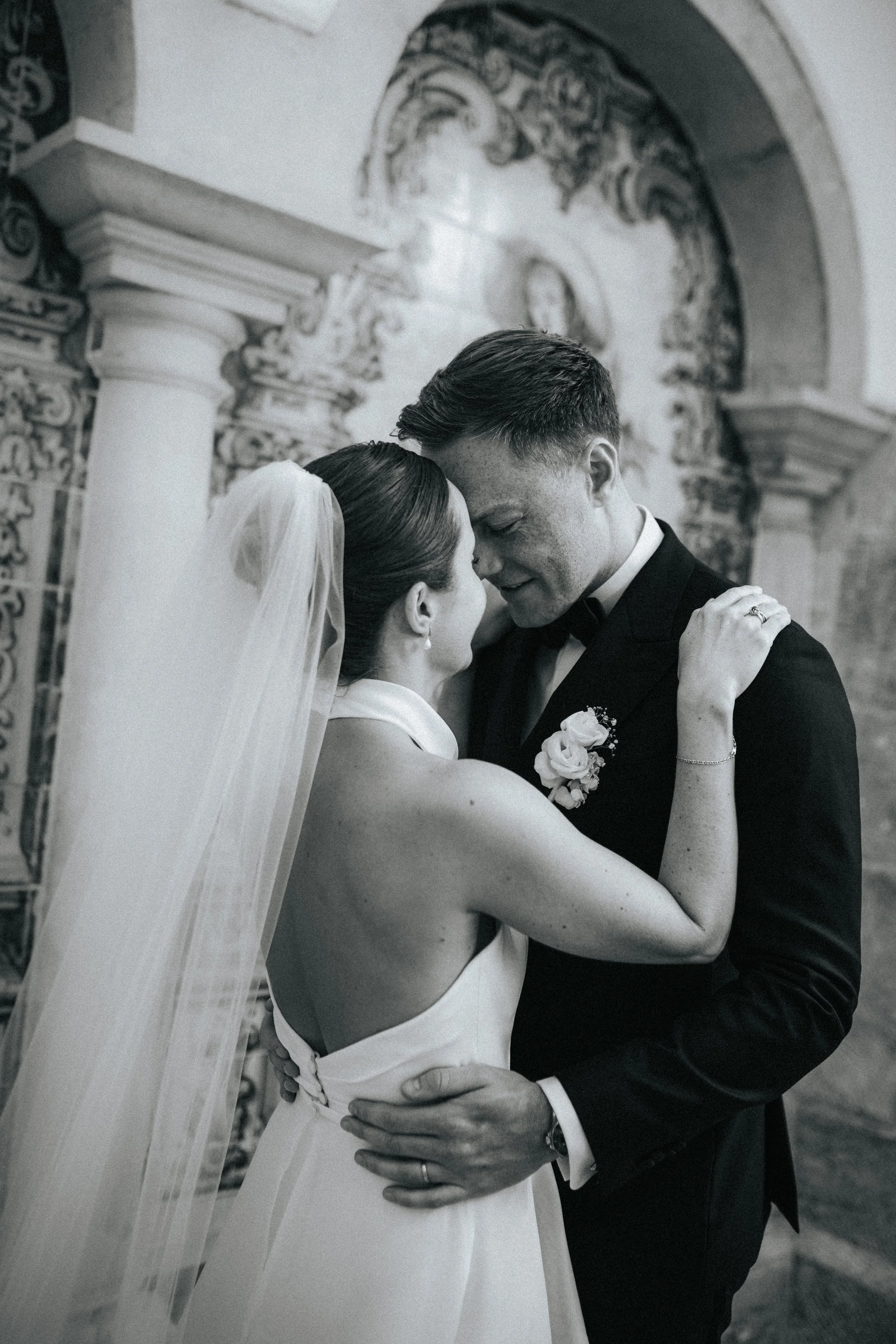 Black and white photo of a bride and groom dancing closely, with the bride wearing a strapless dress and veil, and the groom in a tuxedo, in front of decorative architectural background.