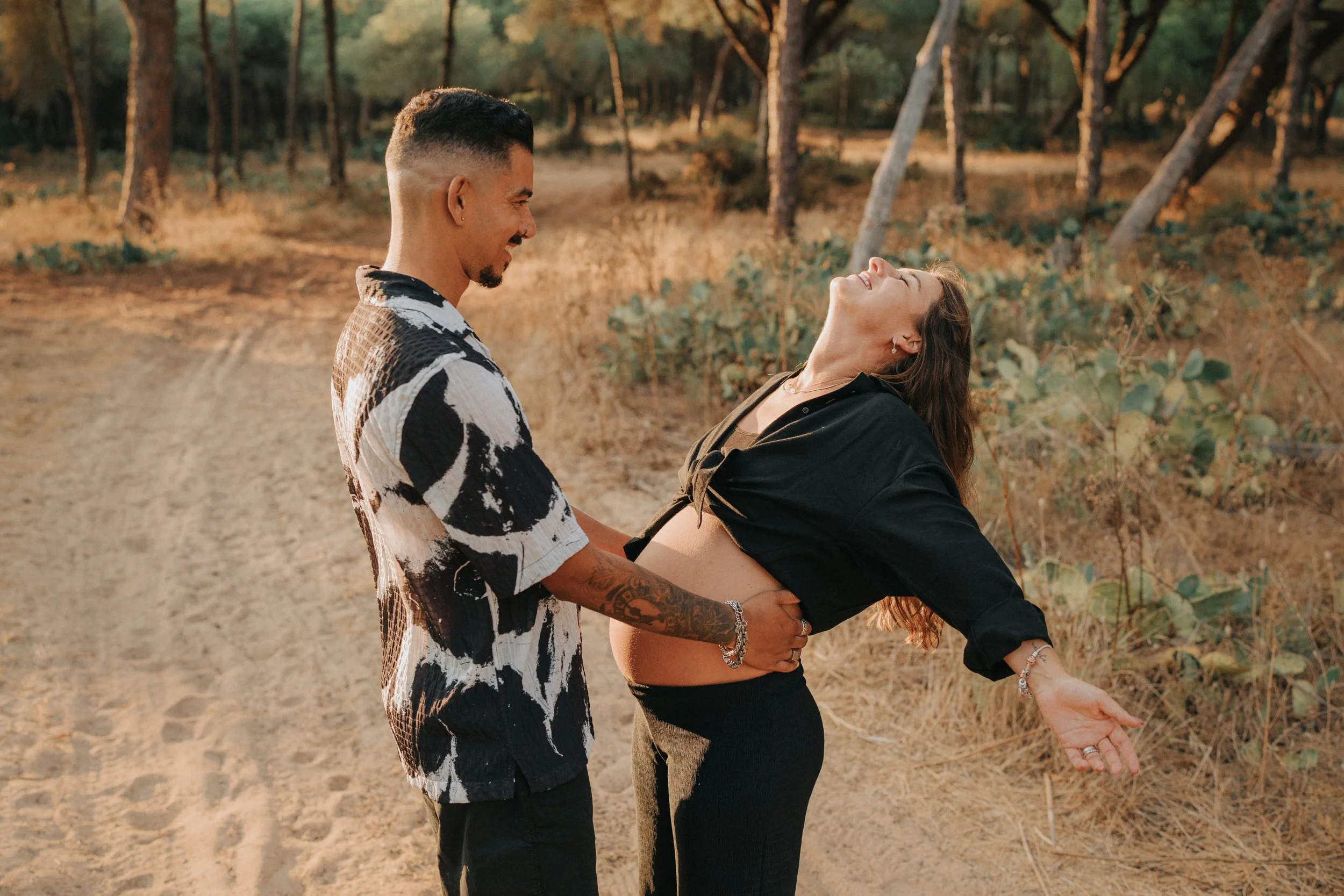 A man and woman are outdoors in a wooded area, sharing a joyful moment with the man holding the woman by her waist, both smiling and laughing.