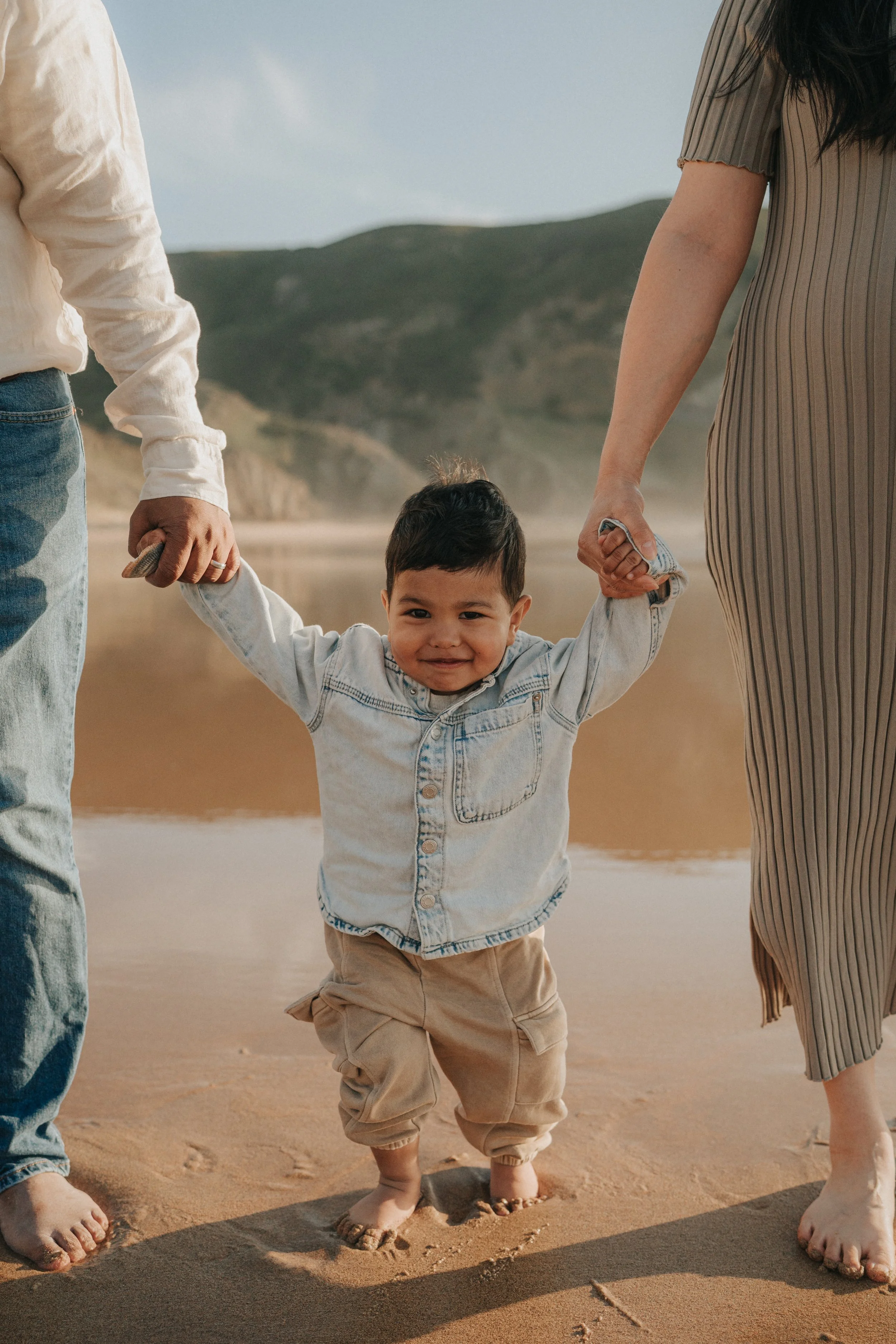 A young boy holding hands with two adults, walking barefoot on the beach, with sand and hills in the background.