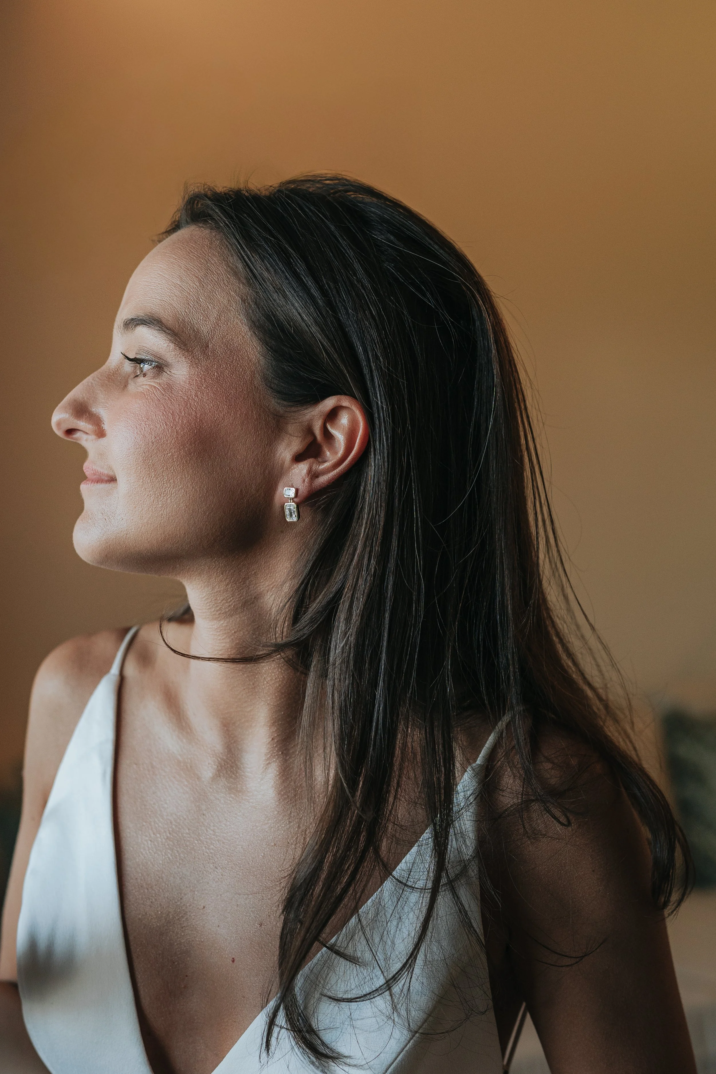 Close-up side profile of a woman with long dark hair, wearing silver earrings and a white sleeveless top against a plain brown background.