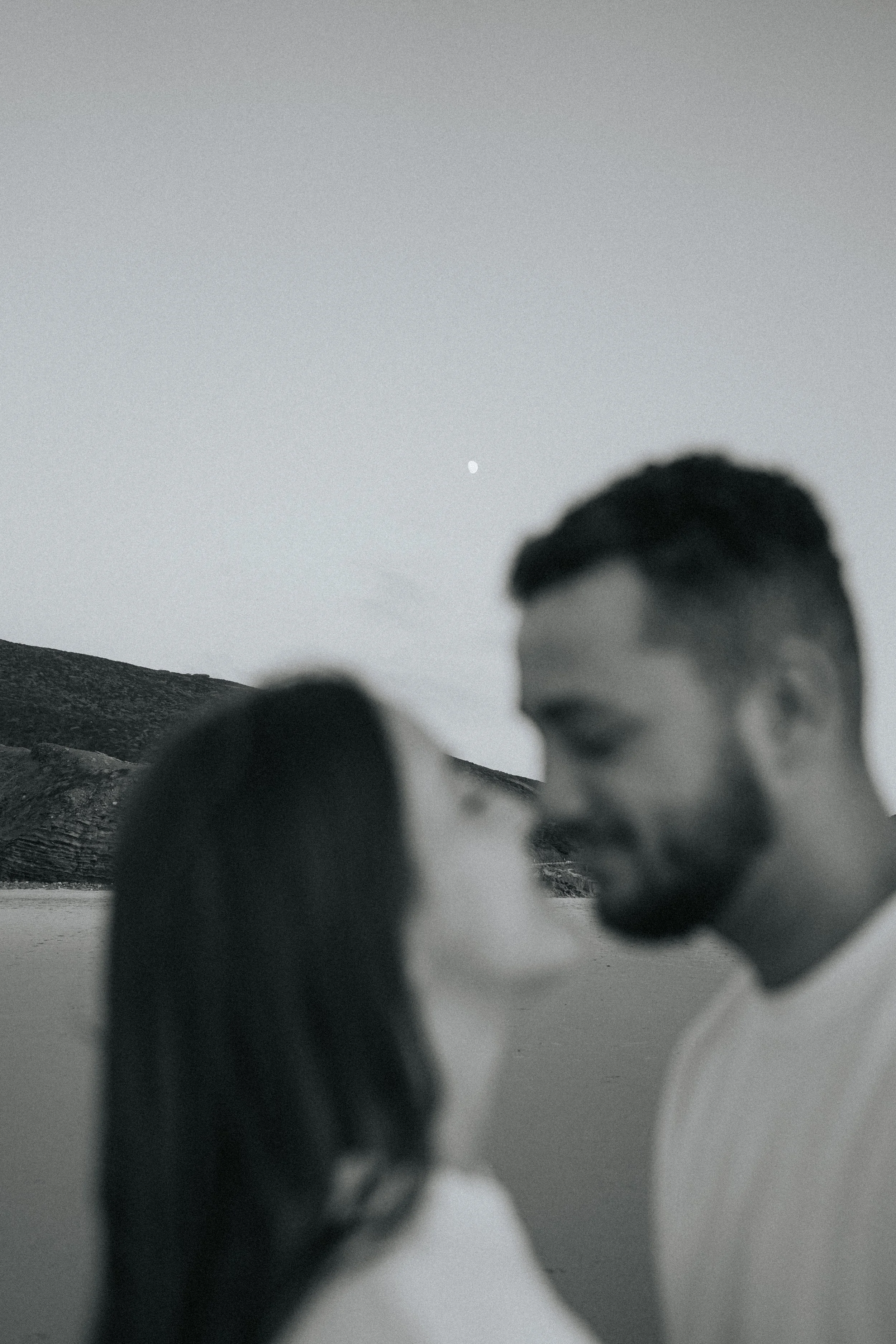 Blurred black and white photo of two people close together with a scenic mountain background and a clear sky with the moon visible.