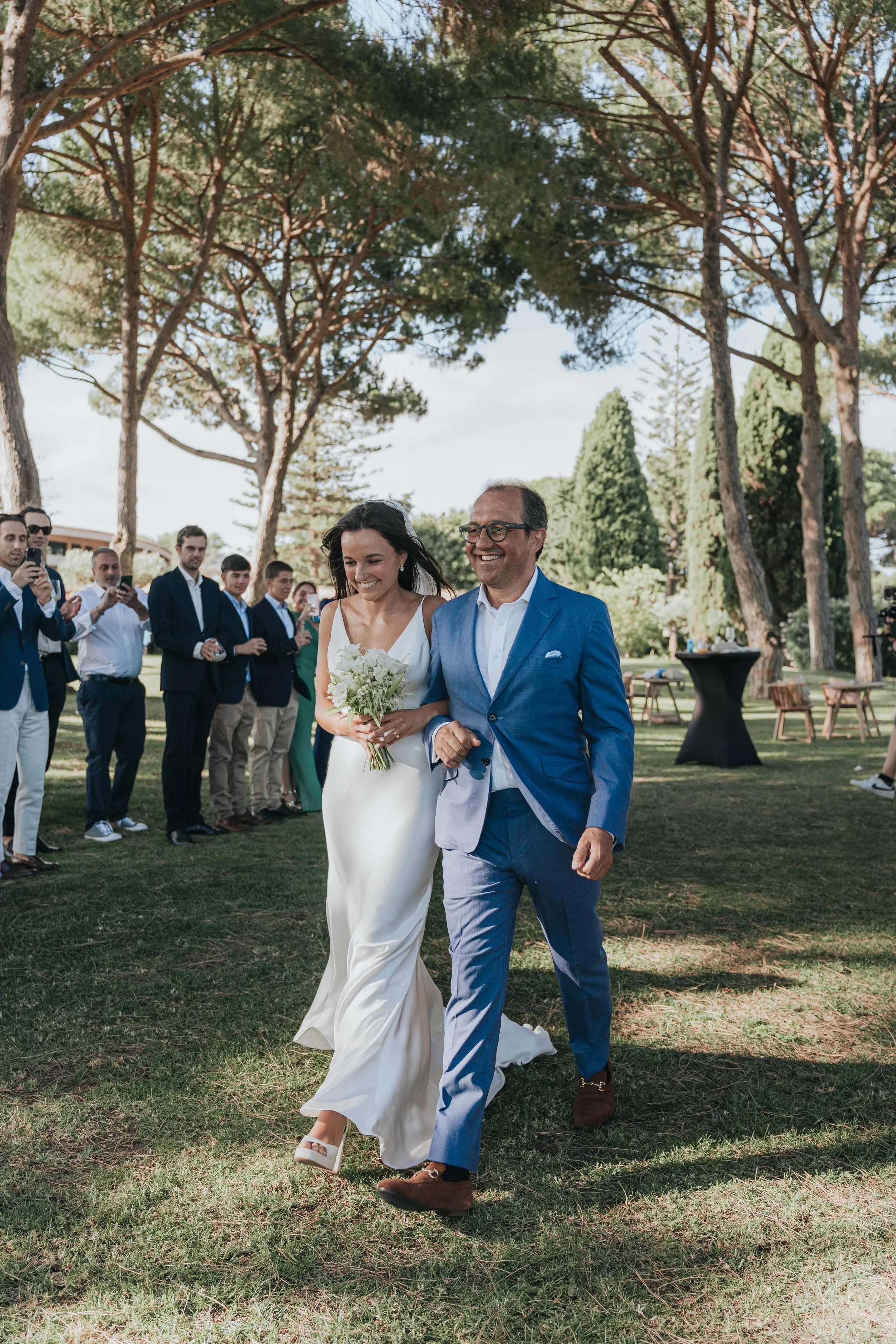 A bride and groom walking outdoors at a wedding reception, surrounded by guests and trees.