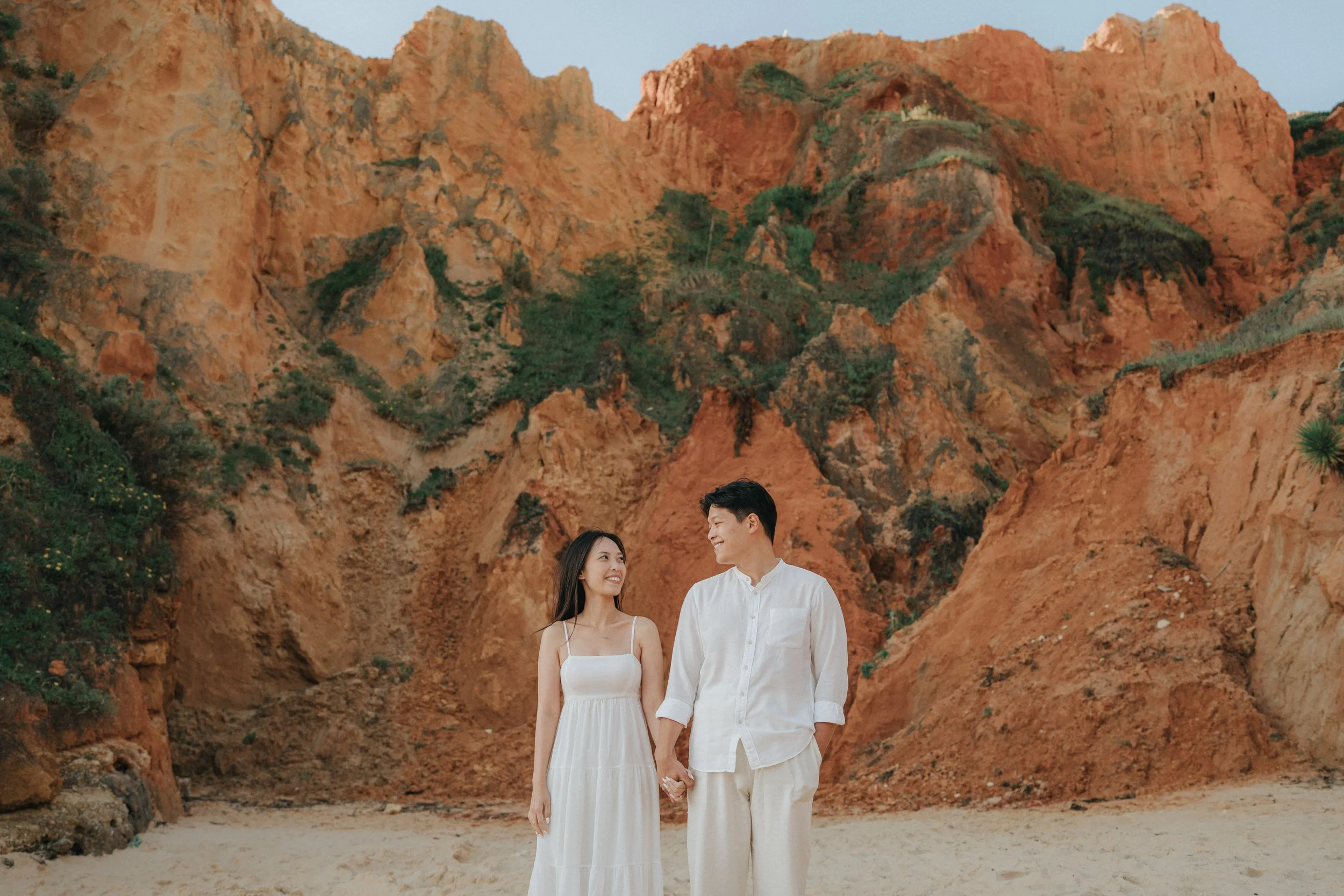 A smiling couple holding hands and walking on a sandy beach with tall orange rock formations in the background.