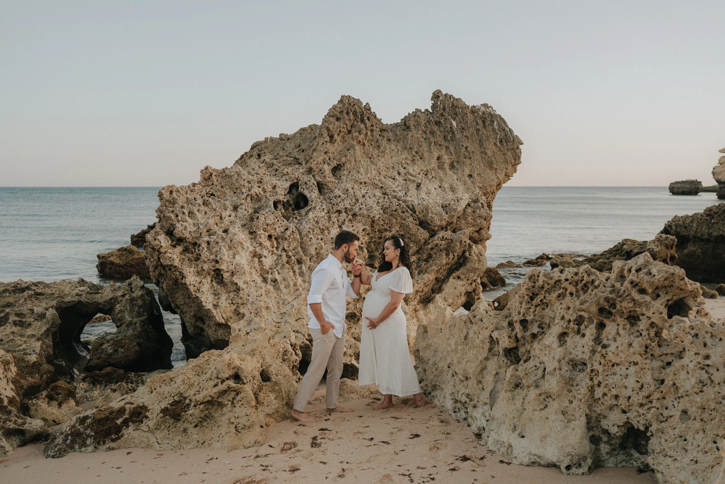 A couple, with the woman visibly pregnant, standing among large rocks at the beach during sunset, holding hands and looking at each other.