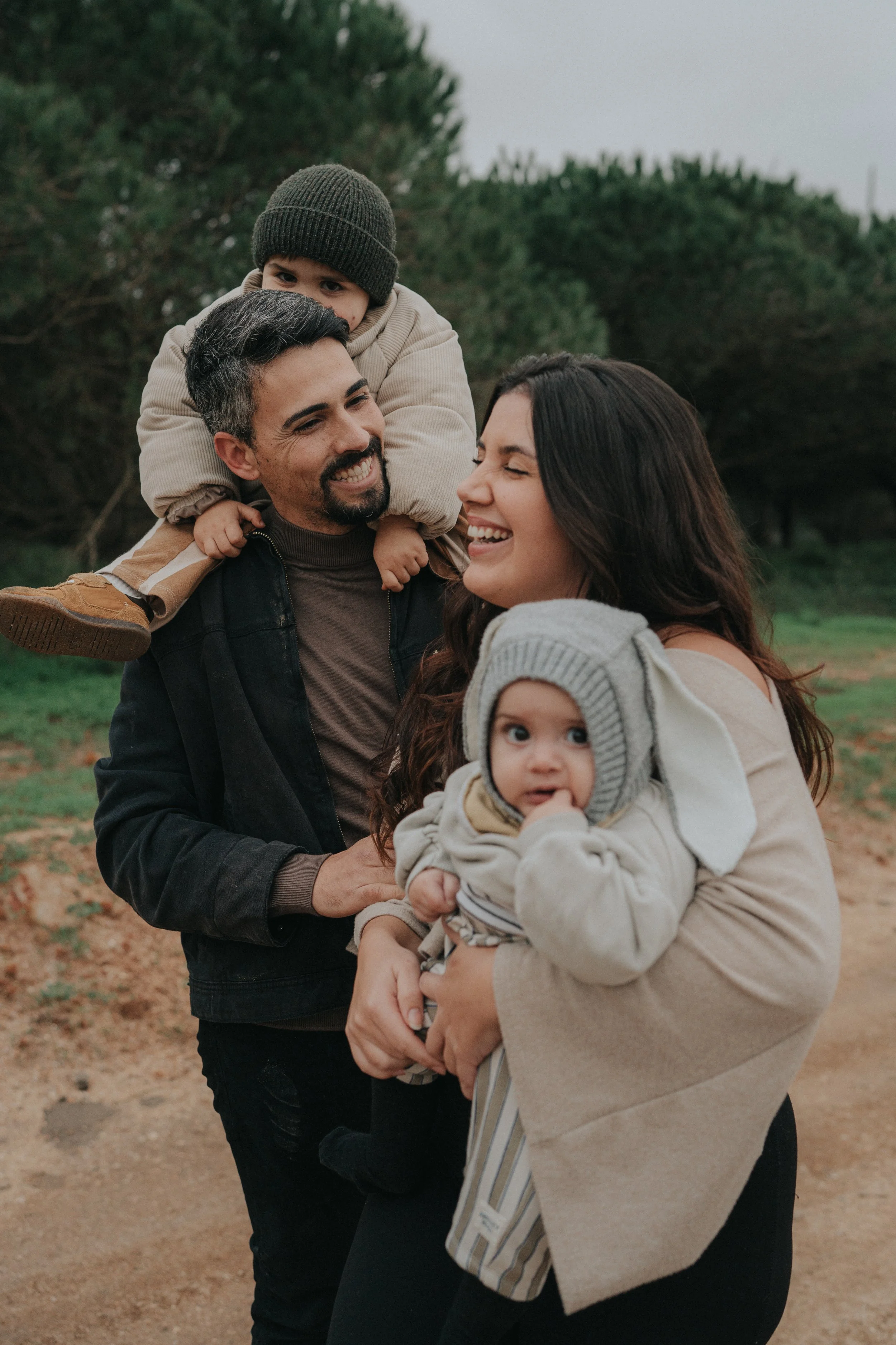 Family of four outdoors, a woman holding a toddler, a man with a child on his shoulders, all smiling and looking at each other.