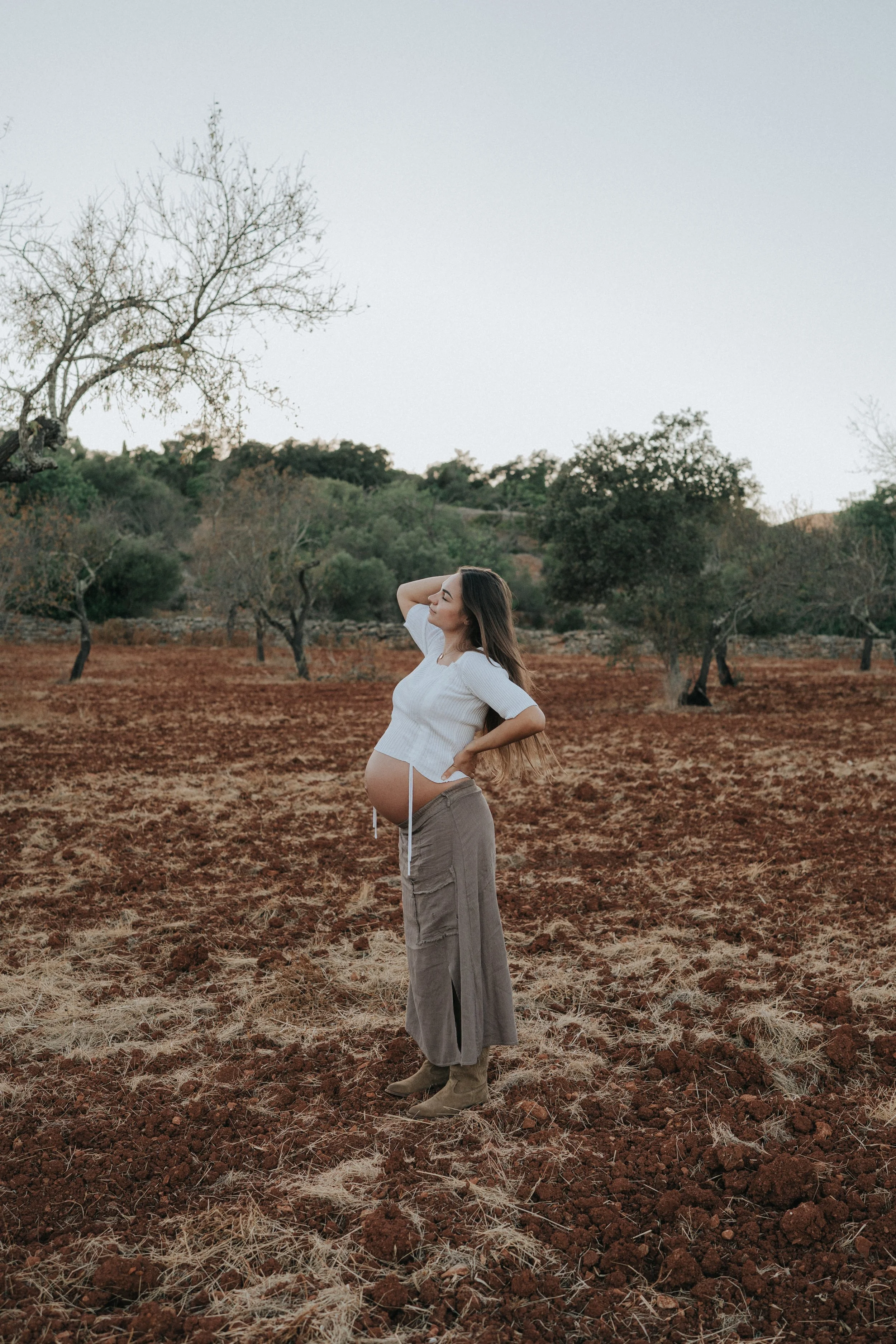 A pregnant woman stands in a rural field with reddish dirt and sparse grass, wearing a white top and loose gray pants, with her head slightly tilted back and one hand on her head, smiling and enjoying the outdoors during daytime, with leafless trees 