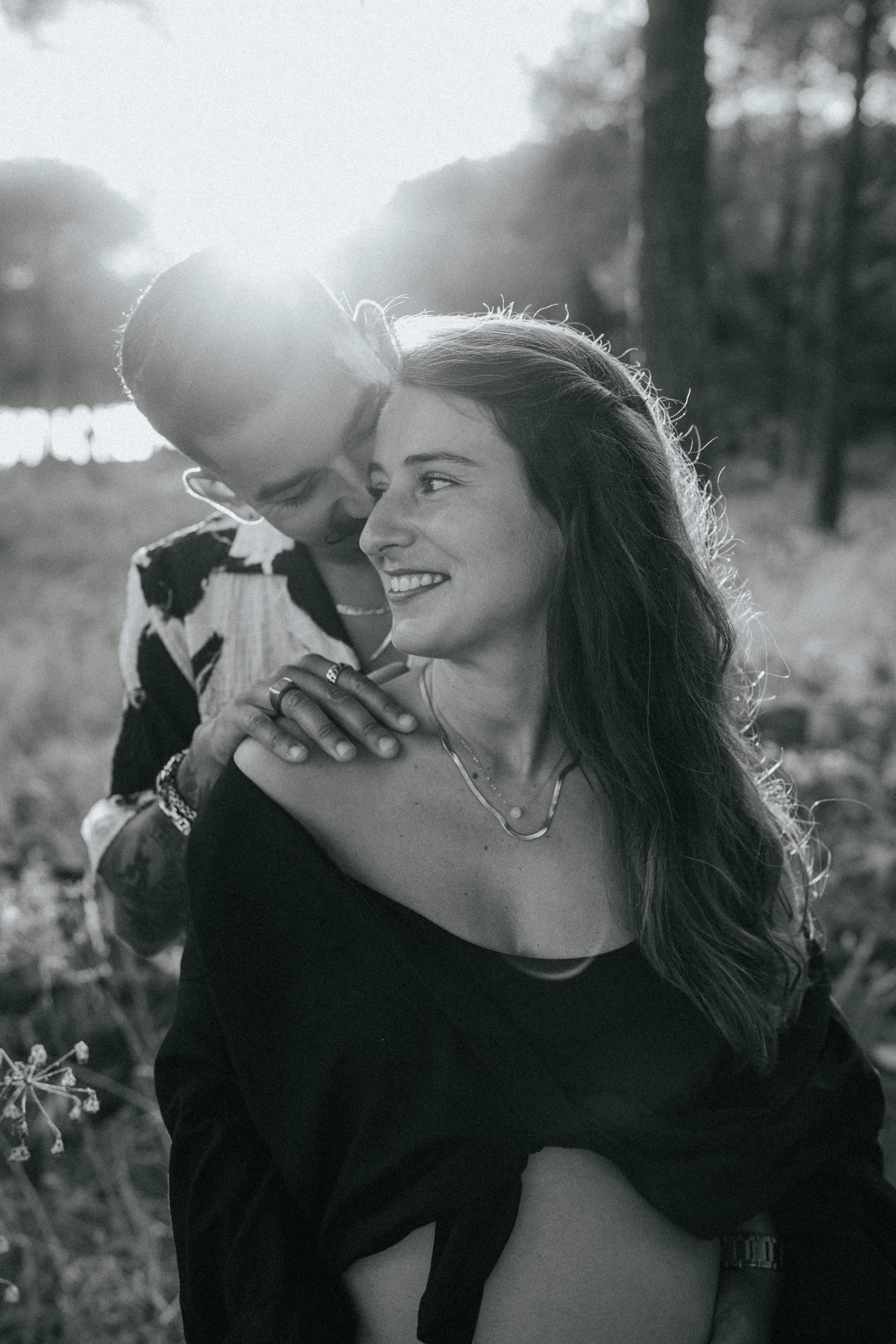A couple hugging outdoors in black and white, with sunlight in the background, in a natural setting with trees and grass.