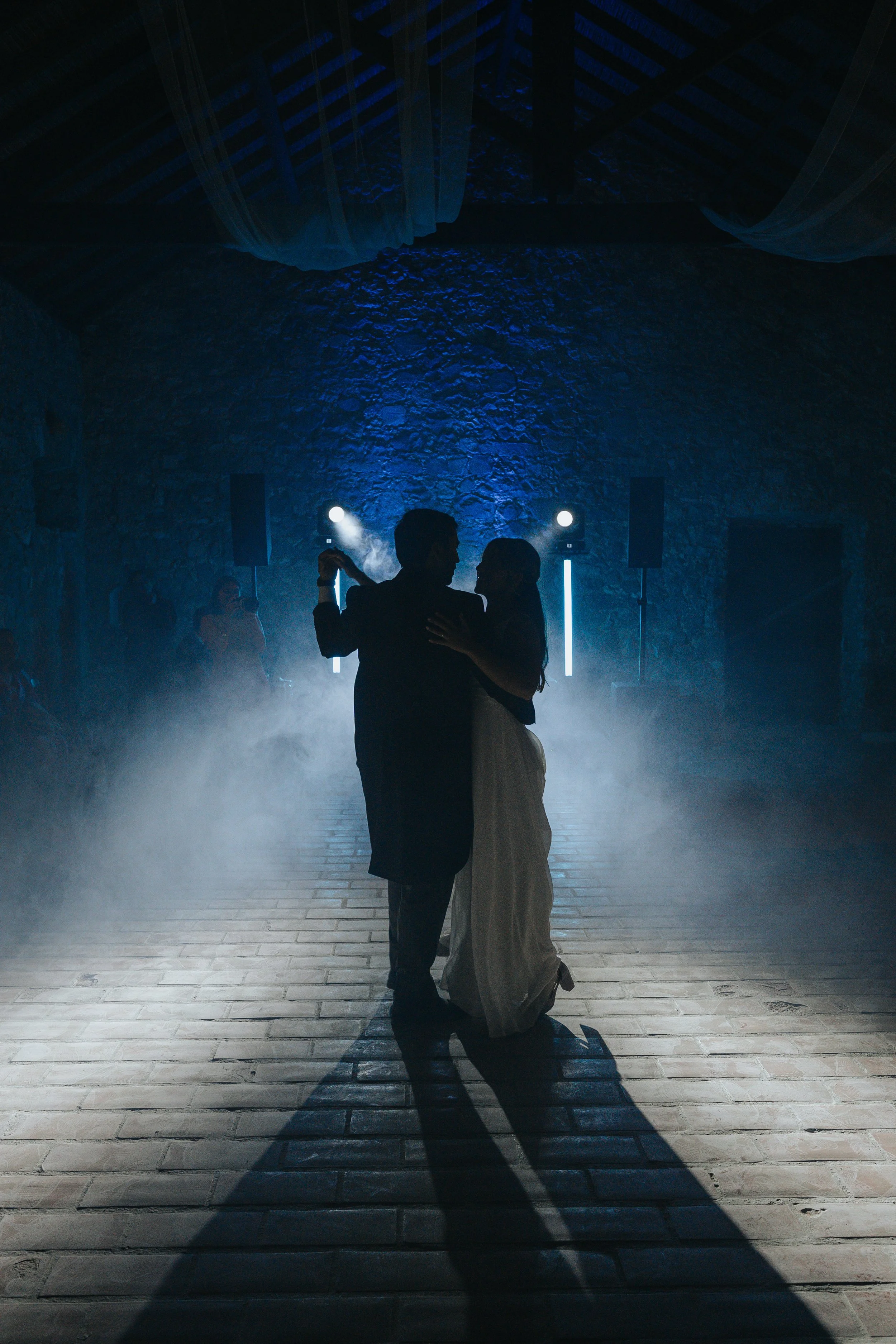 A silhouetted couple sharing a dance in a dimly lit, atmospheric venue with a stone wall background and fog or smoke at their feet, illuminated by blue and white stage lights.