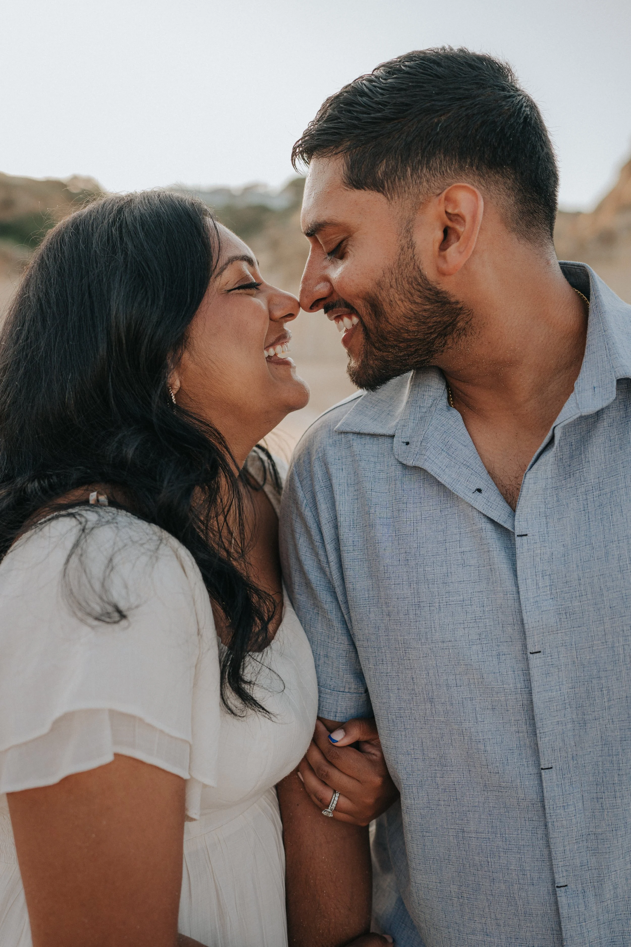 A couple smiling with foreheads touching, holding hands, outdoors in daylight.