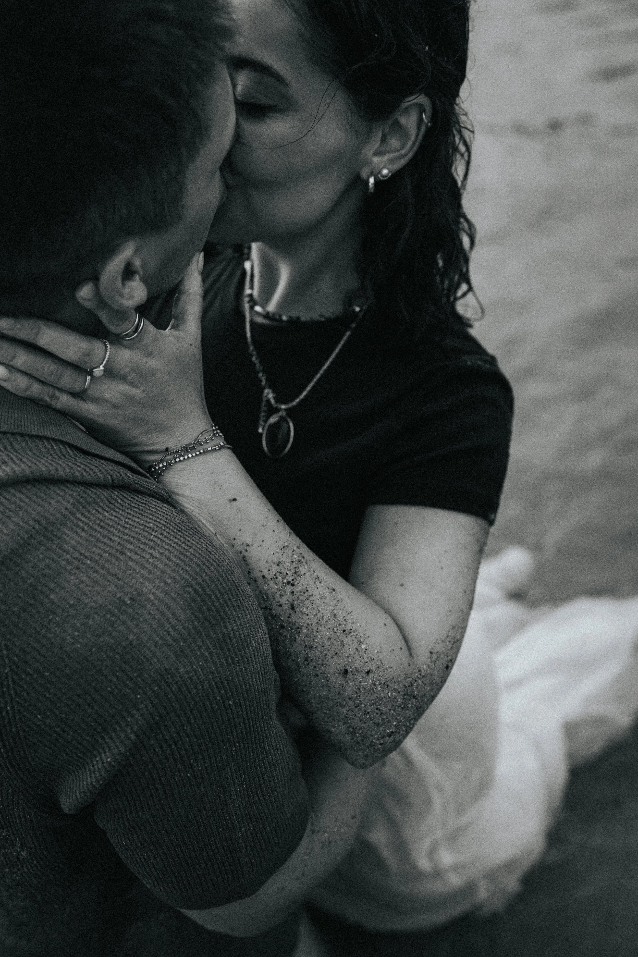 A black and white photo of a man and woman embracing on the beach, sharing a kiss. The woman's arm and back are visible, and sand is on her arm. The woman is wearing jewelry and a dark shirt. The man's face is partly obscured.