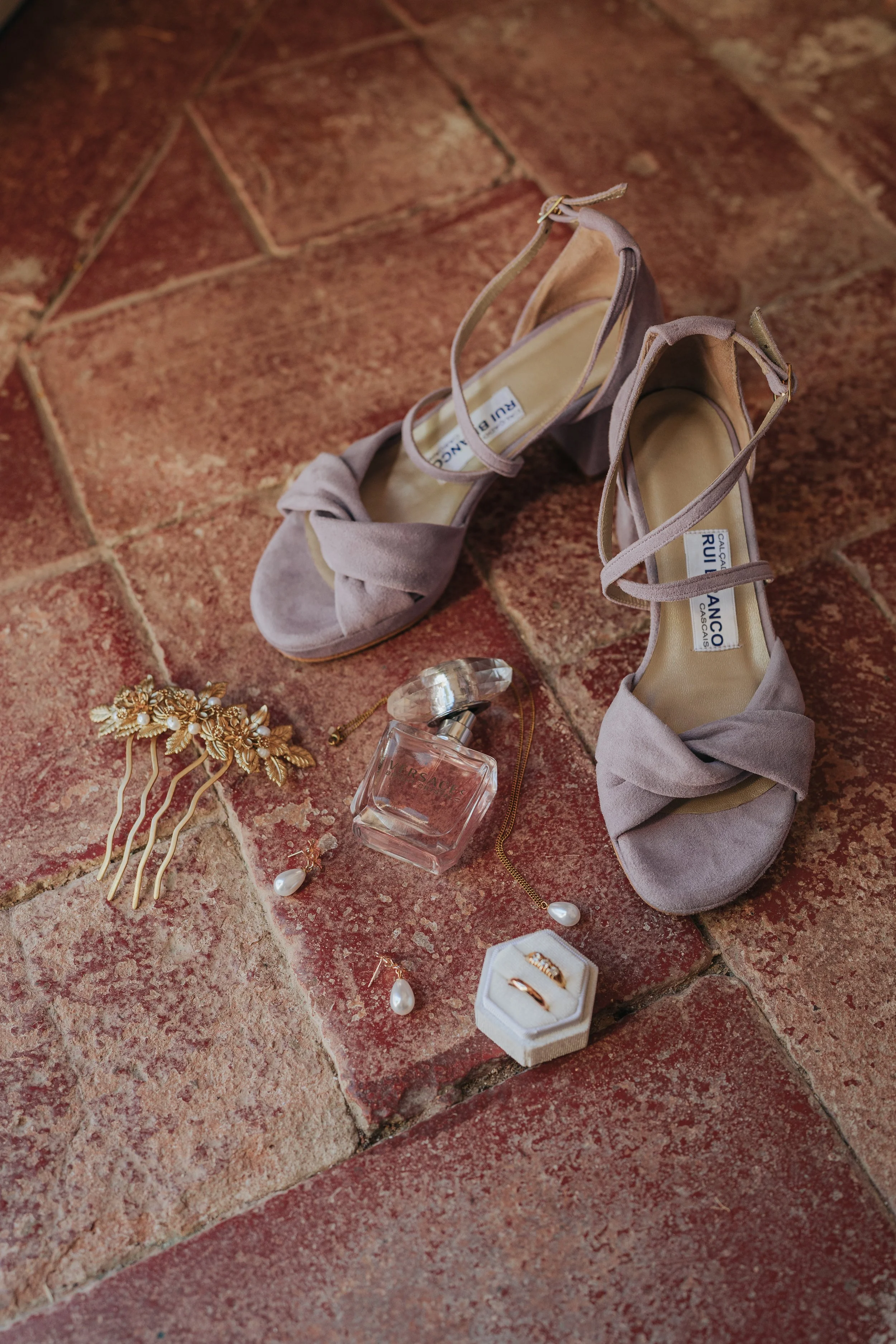 A pair of gray high-heeled shoes with ankle straps placed on a brick floor. Surrounding the shoes are jewelry items including a pearl ring in a white box, a pearl necklace, a gold hair accessory with leaves and flowers, a pair of pearl earrings, and 