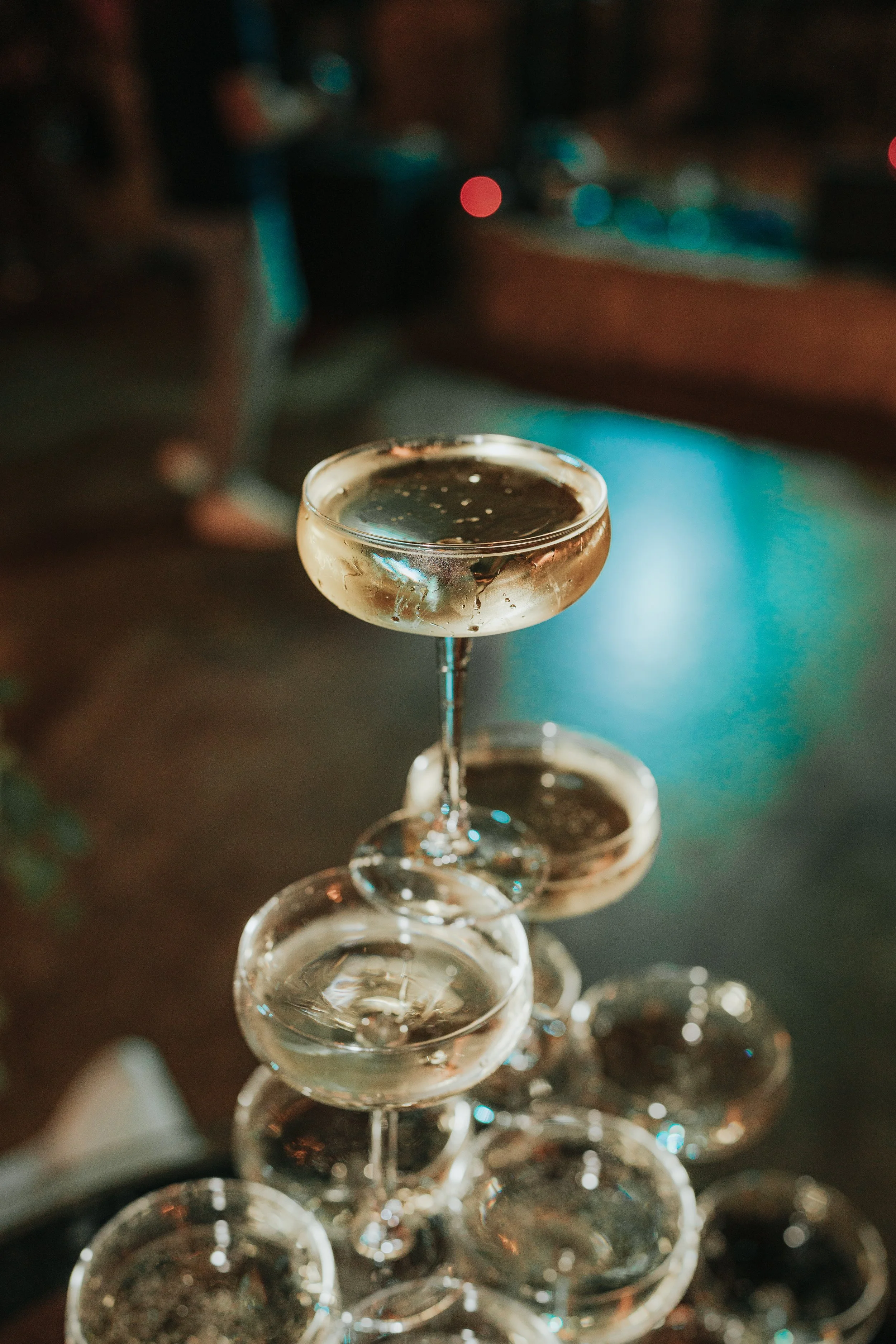 A tower of clear glass coupe glasses filled with a dark liquid, arranged in a cone shape in a dimly lit setting with colorful bokeh lights in the background.