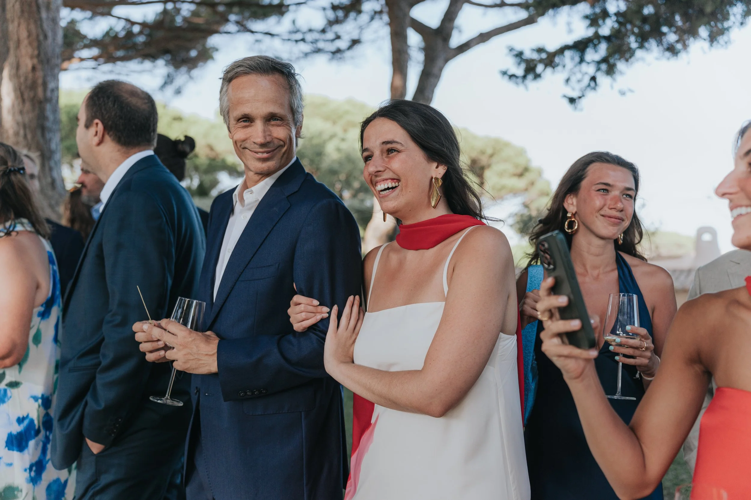 Group of people attending outdoor social gathering, smiling and dressed in semi-formal attire, holding glasses of champagne or wine, under large trees with clear sky.