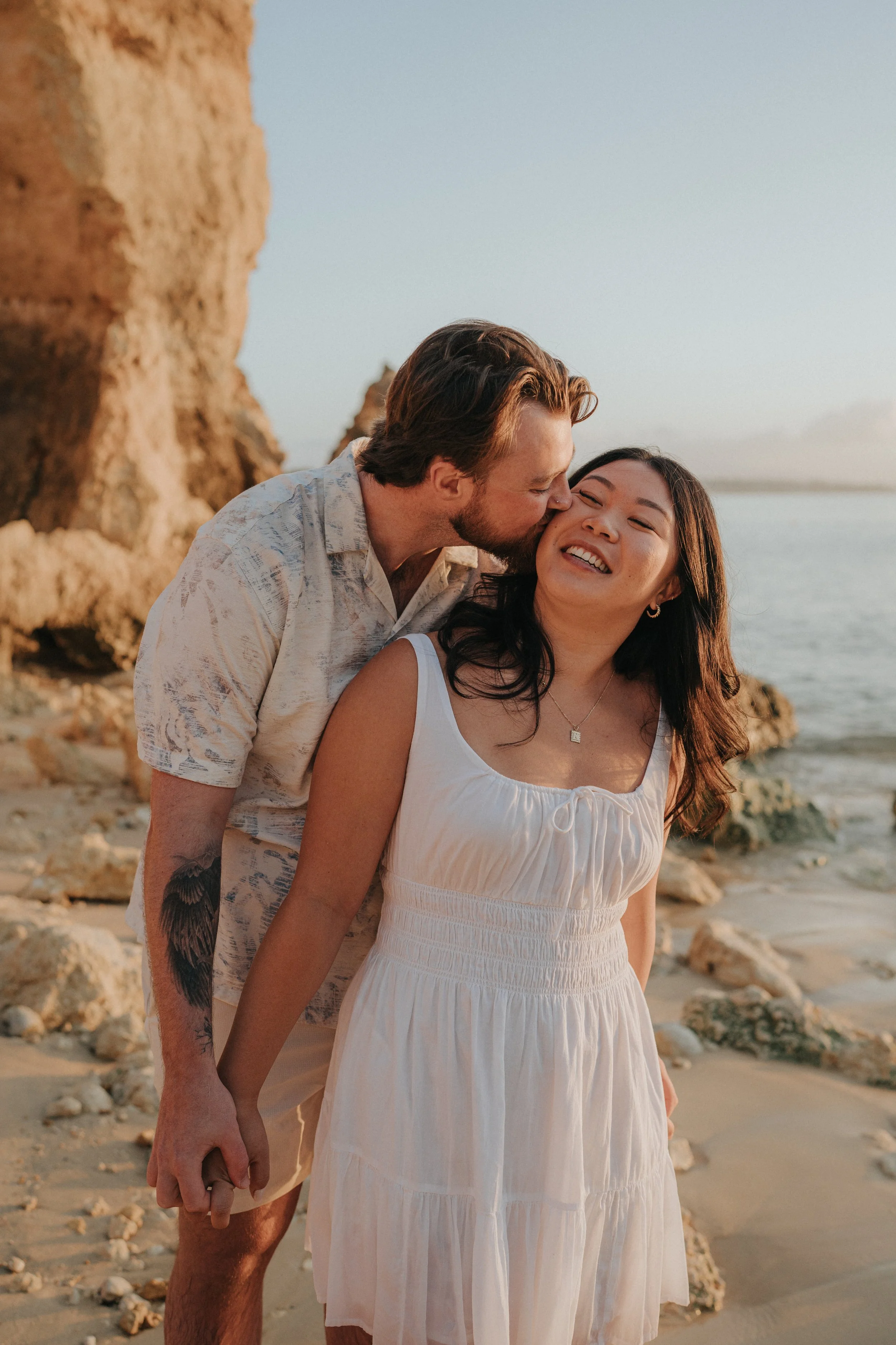 A couple at the beach during sunset, holding hands and smiling. The man is kissing the woman on her cheek, both dressed in light summer clothes, with rocks and the ocean in the background.