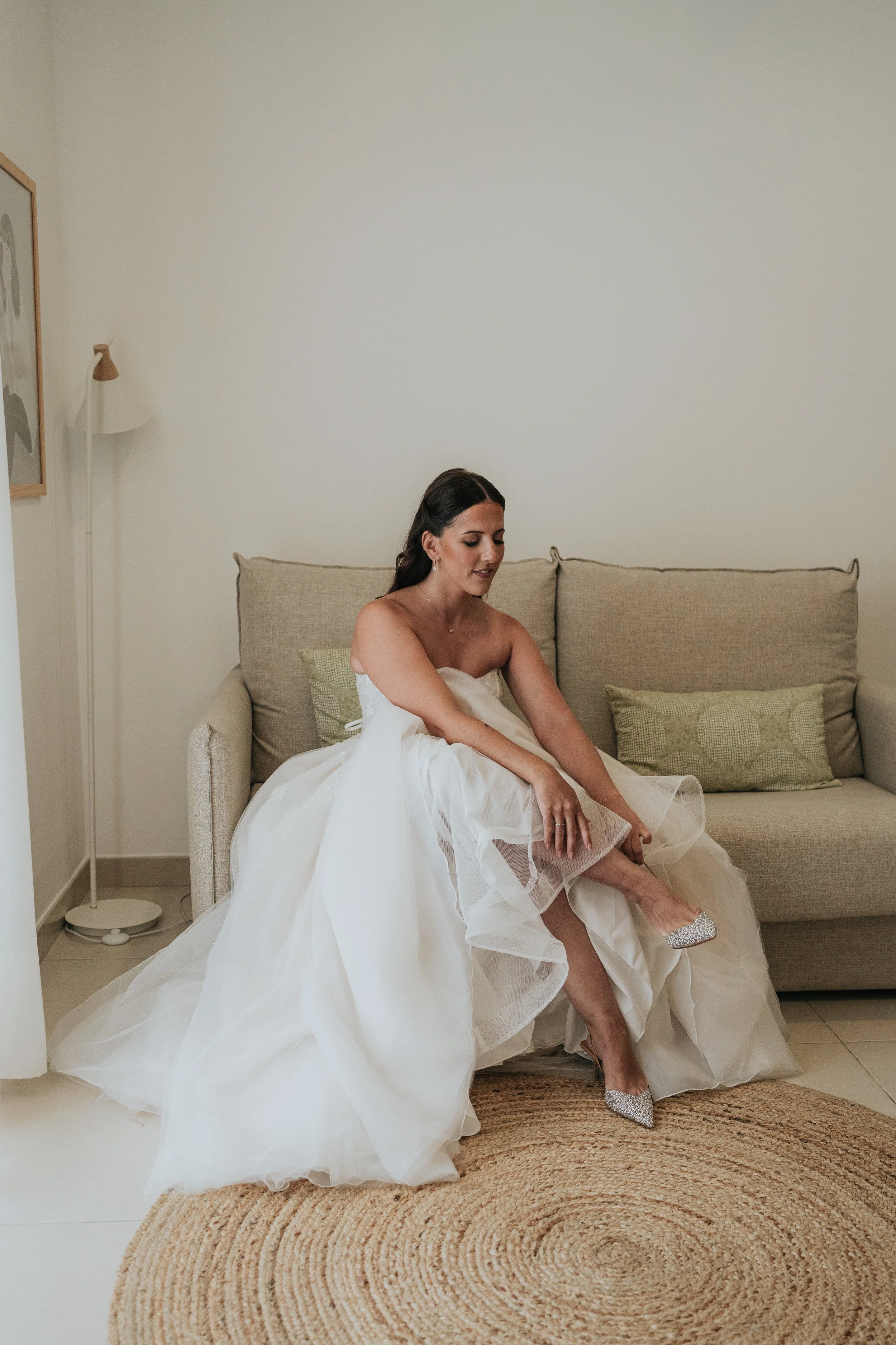 A woman in a wedding dress sitting on a beige sofa in a room, adjusting her shoe.