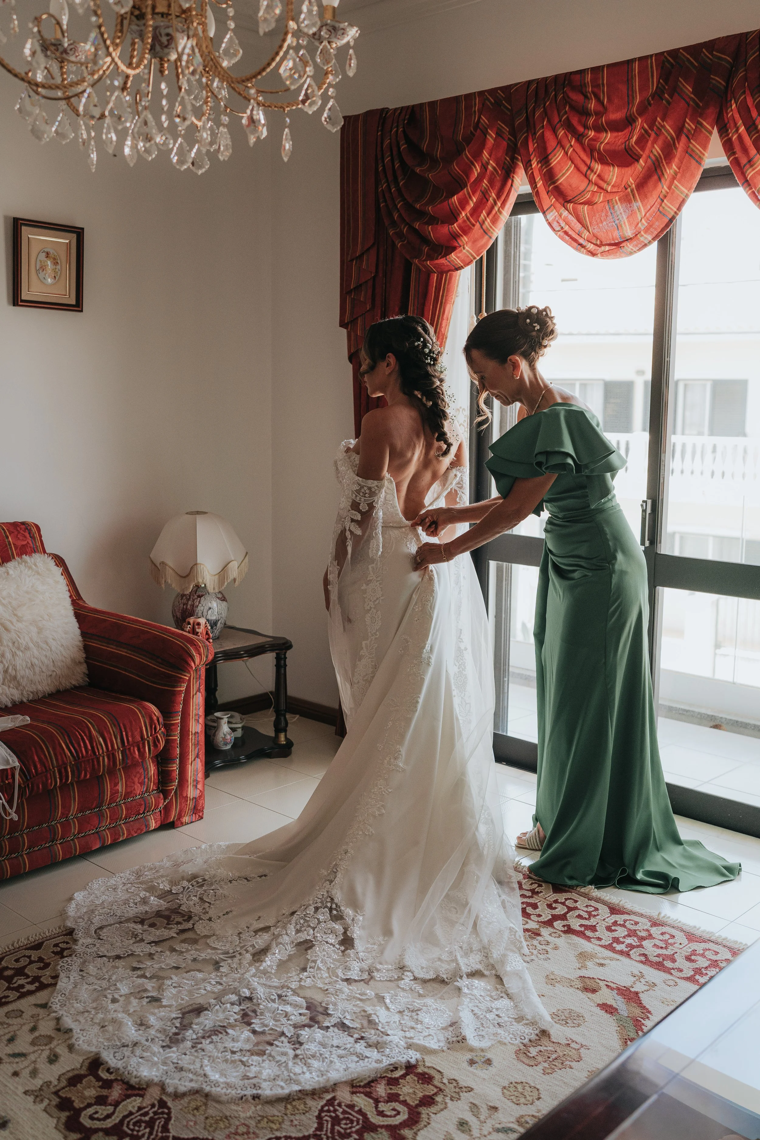 A bride in a white wedding dress standing indoors near a large window while another woman, likely her bridesmaid or mother, helps her with the dress. The room has a chandelier, patterned curtains, a sofa, and a rug.