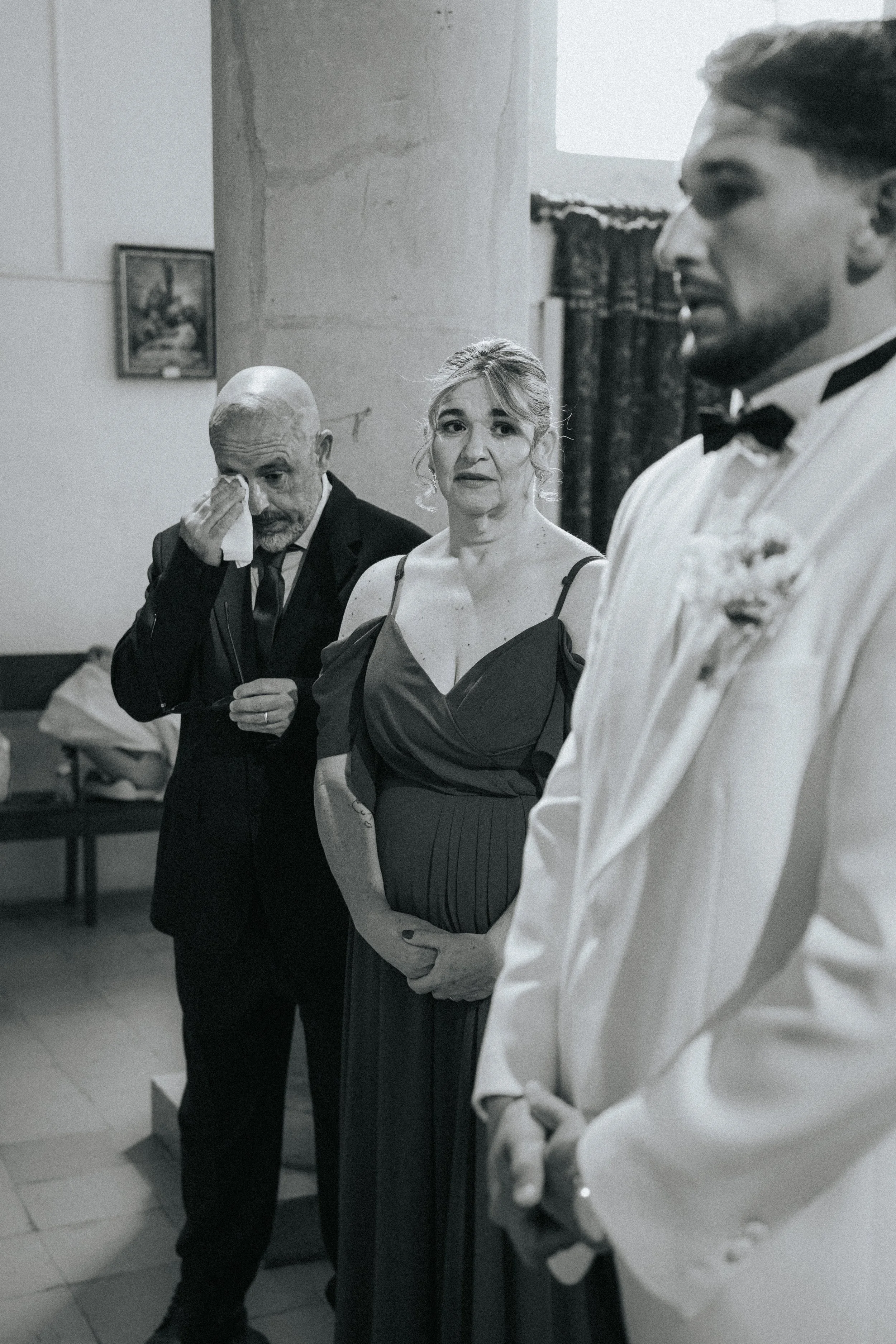 Three people standing in a room, dressed formally. One man on the left is wiping his eye with a tissue. The woman in the center is looking at the camera with a serious expression. The man on the right is in profile, wearing a tuxedo, with his hands c