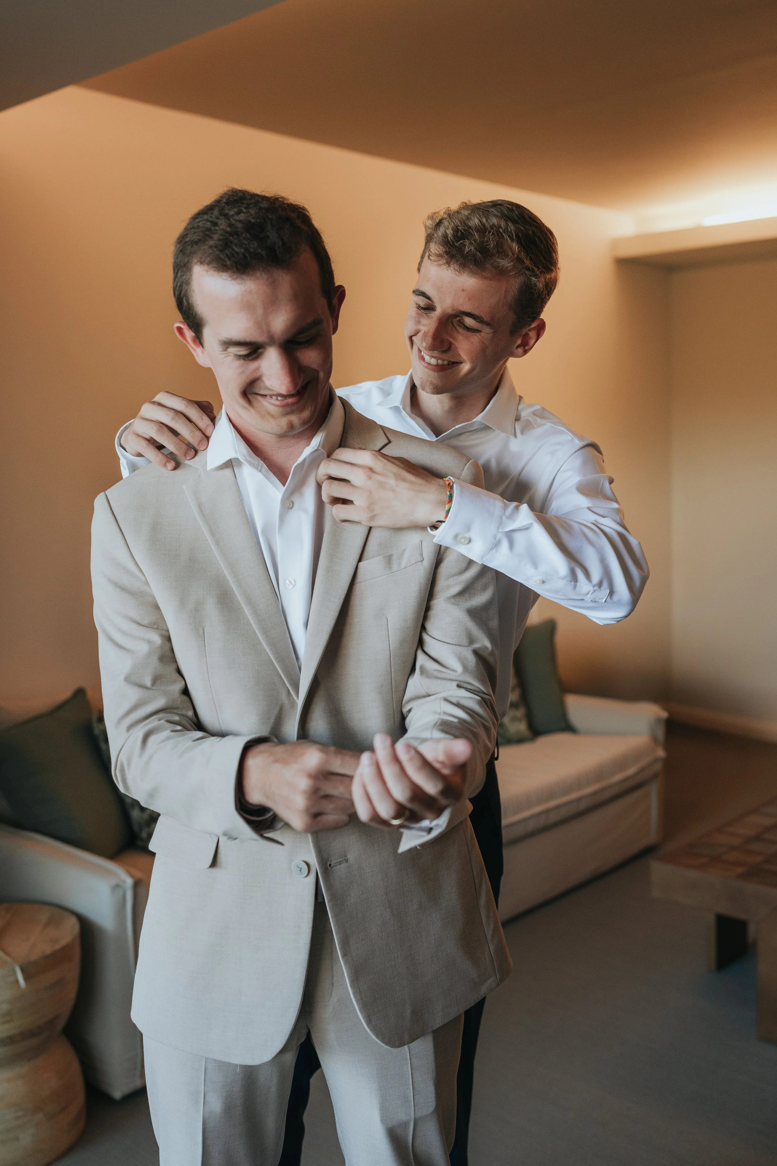 Two young men in formal attire, one adjusting the other's suit, smiling in a warmly lit room.