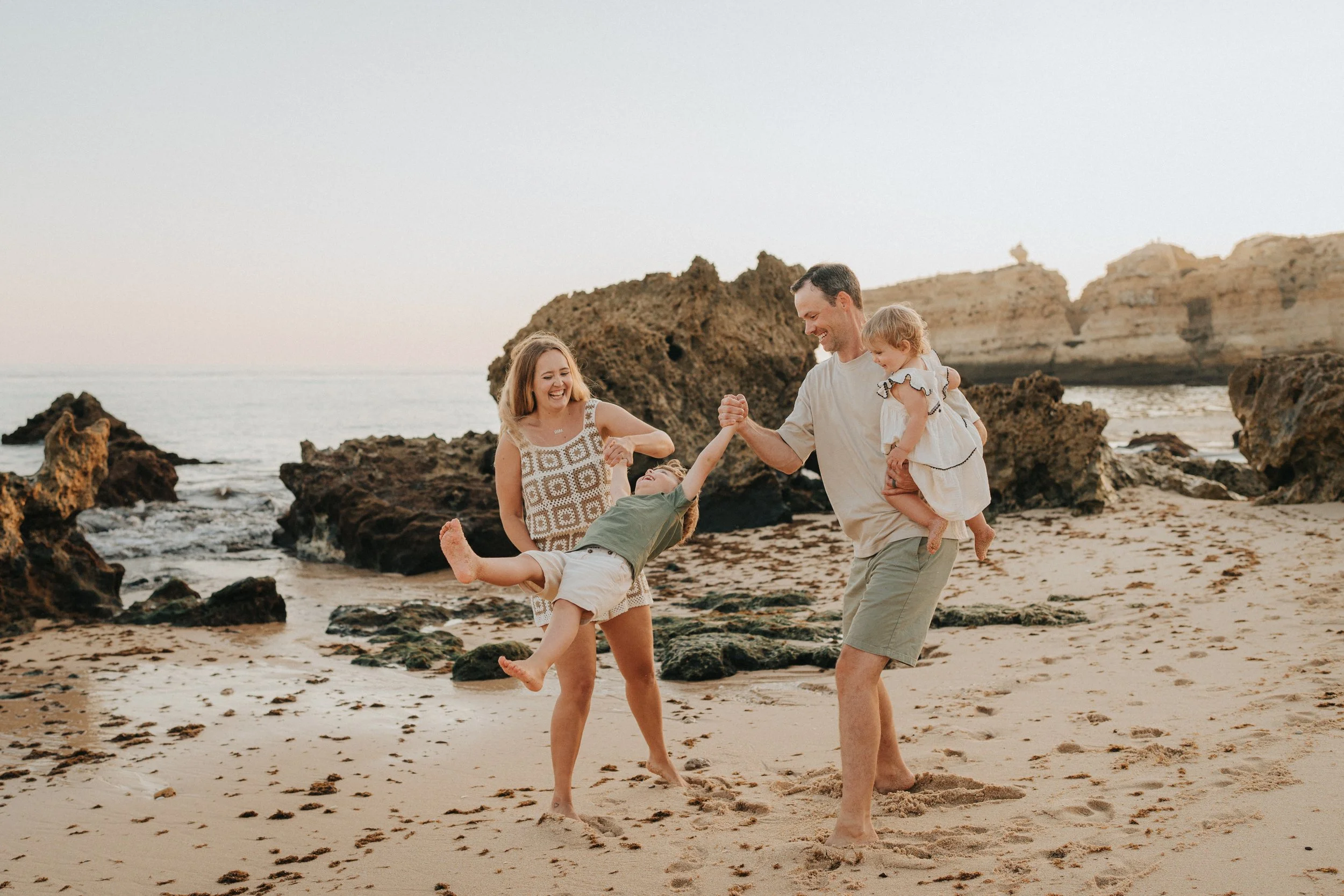 A family of four playing and laughing on the beach near rocky formations at sunset.