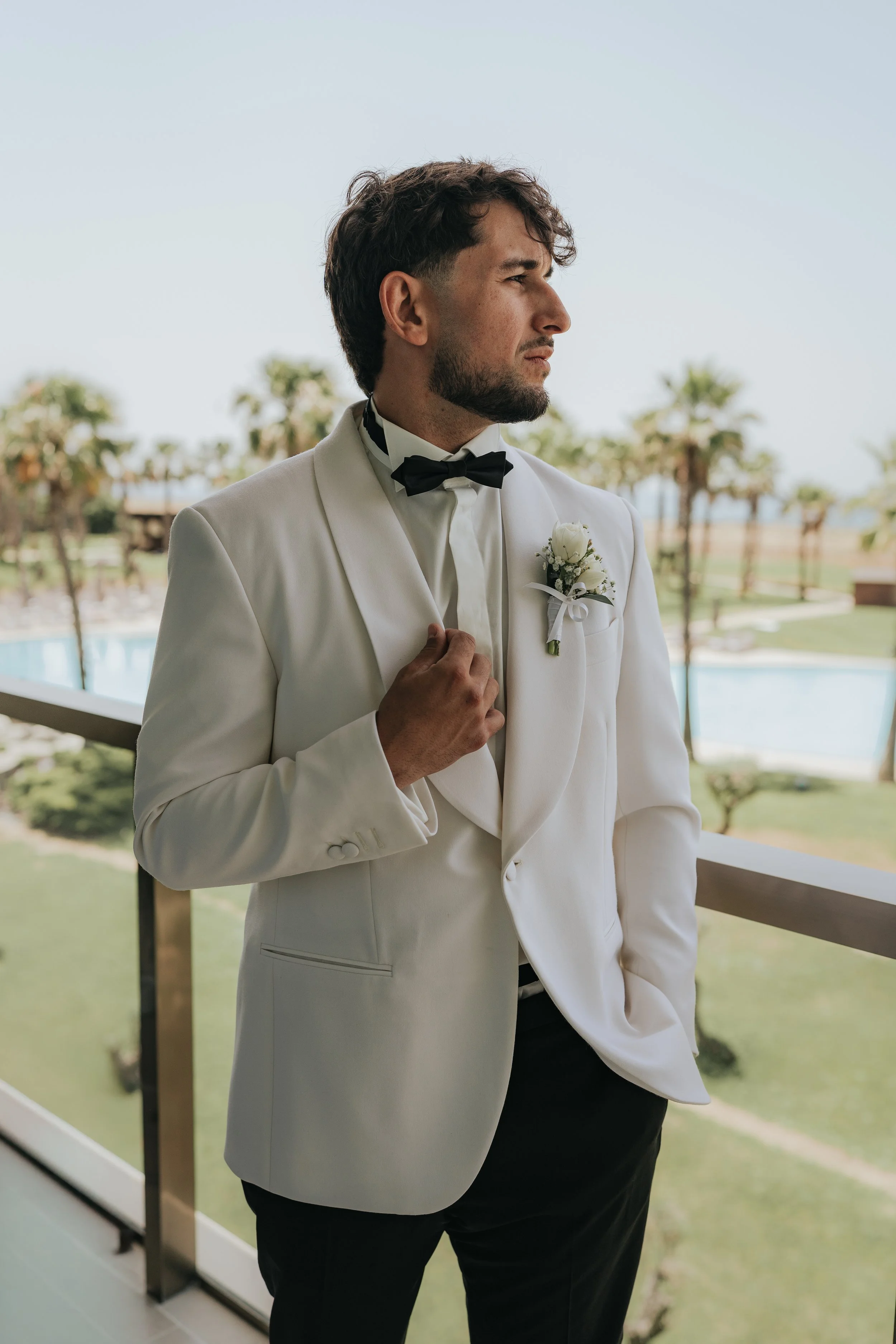 A man dressed in a white tuxedo with a black bow tie, standing on a balcony. He has a boutonniere on his lapel, and is looking to the right with a serious expression. Palm trees and a swimming pool are visible in the background.