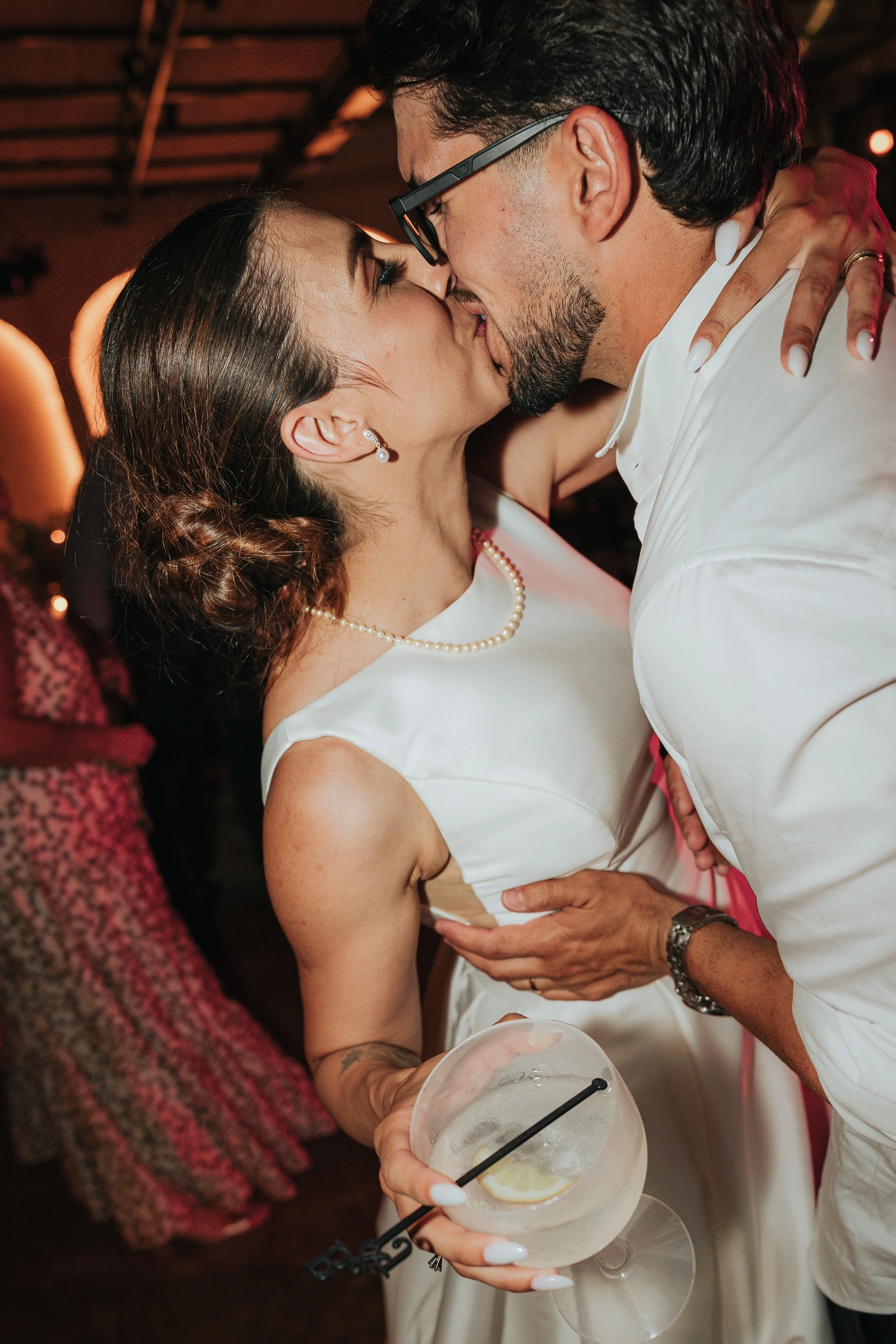 A couple sharing a kiss at a celebration or party. The woman holds a cocktail glass with a lemon slice and straw, wearing pearl earrings, a necklace, and a sleeveless white dress. The man wears glasses and a white shirt.