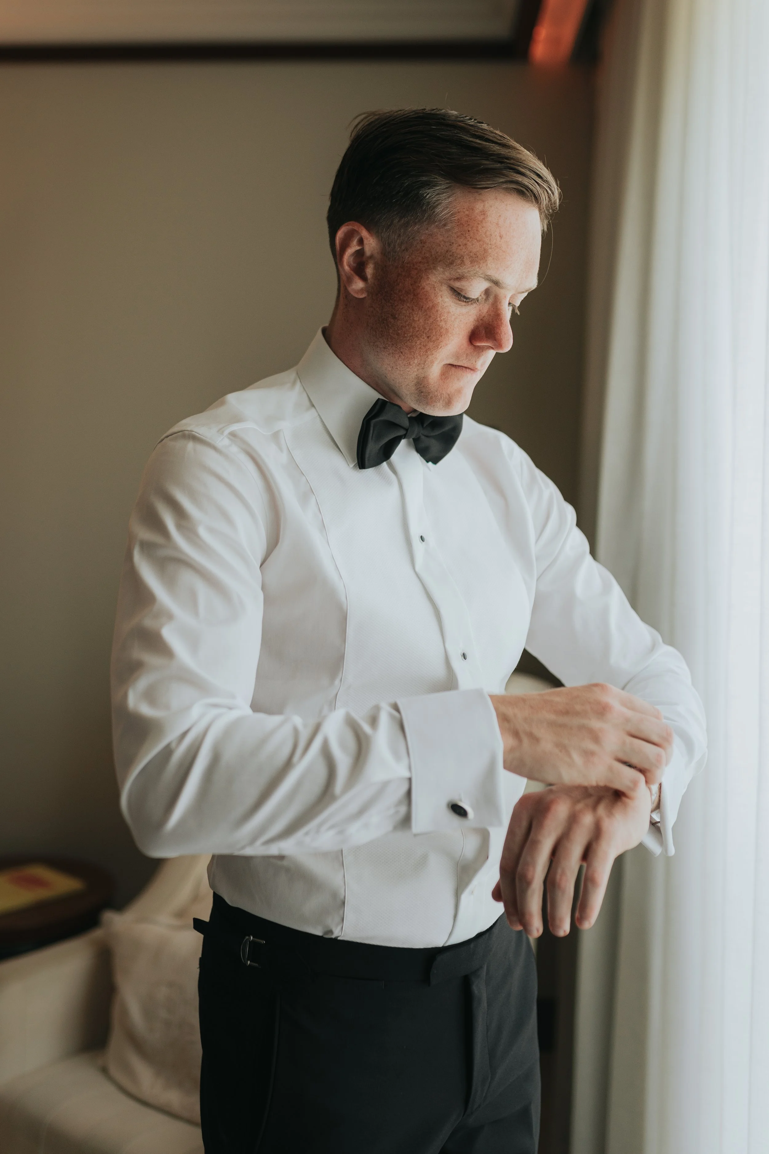 A man in a white dress shirt with black buttons and a black bow tie, adjusting his cufflinks in a room with beige walls and a window with curtains.