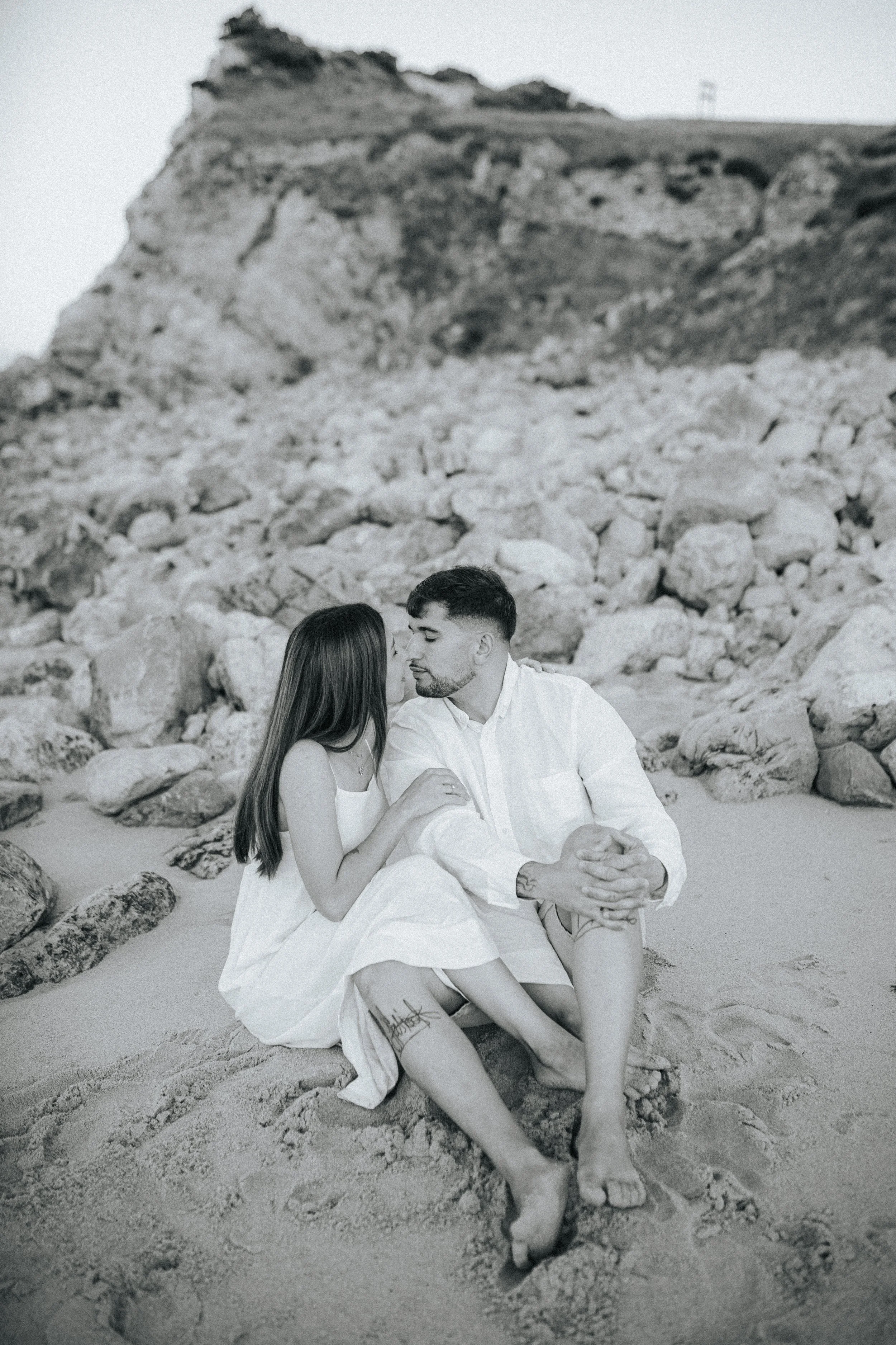 A black and white photo of a couple sitting on the sand at the beach, close to a rocky hillside, with the woman holding the man's hand and facing each other.