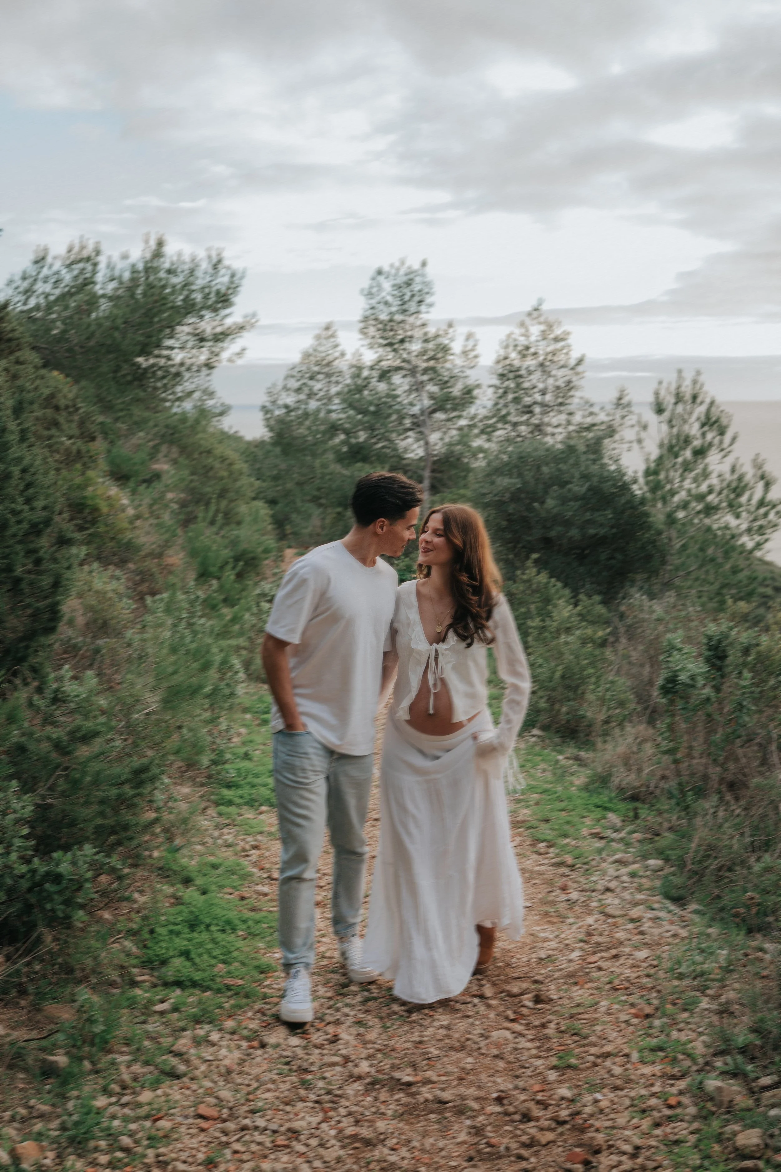A young couple walking on a trail through a wooded area with trees, rocks, and cloudy sky overhead.