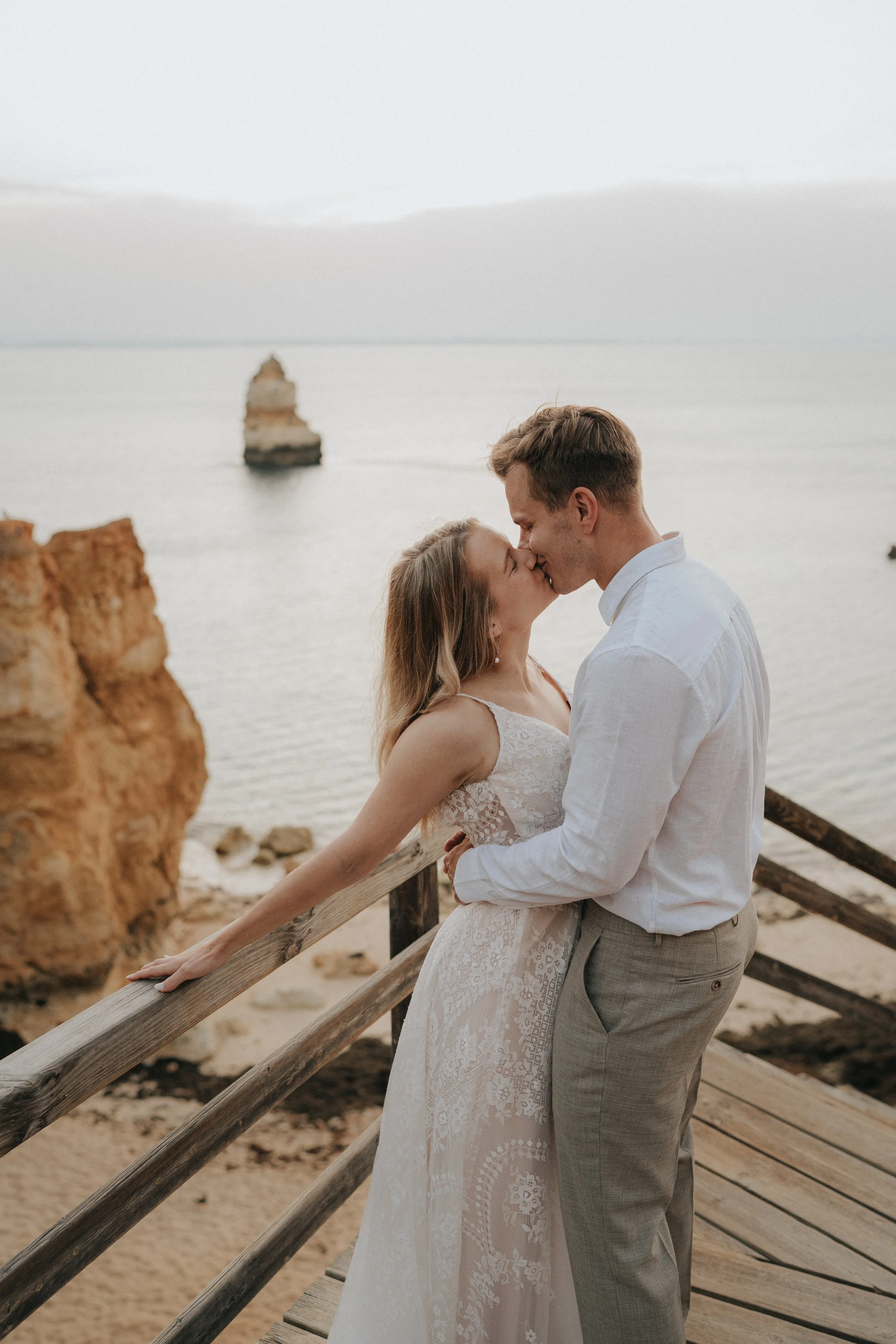 A couple dressed in wedding attire sharing a kiss by the ocean, with a large rock formation in the water and a wooden railing on a beach.