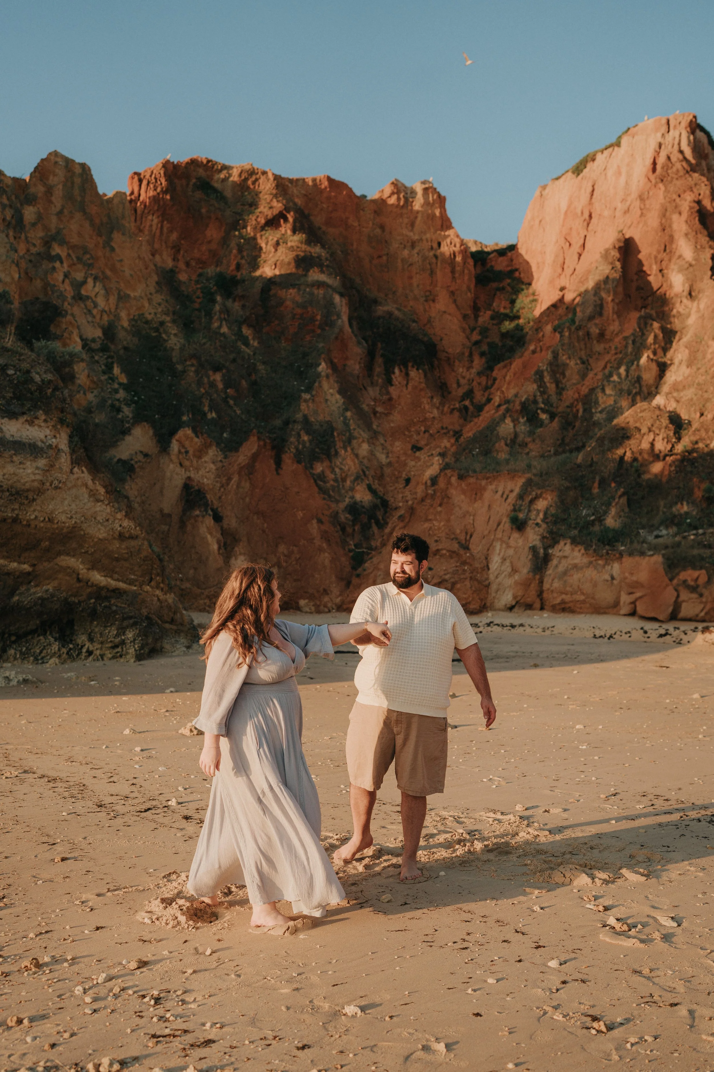 A couple walking along a sandy beach with large rocky cliffs in the background and a blue sky. The woman is wearing a long, light-colored dress and has curly hair, while the man is wearing a white shirt and brown shorts, with facial hair. The woman i