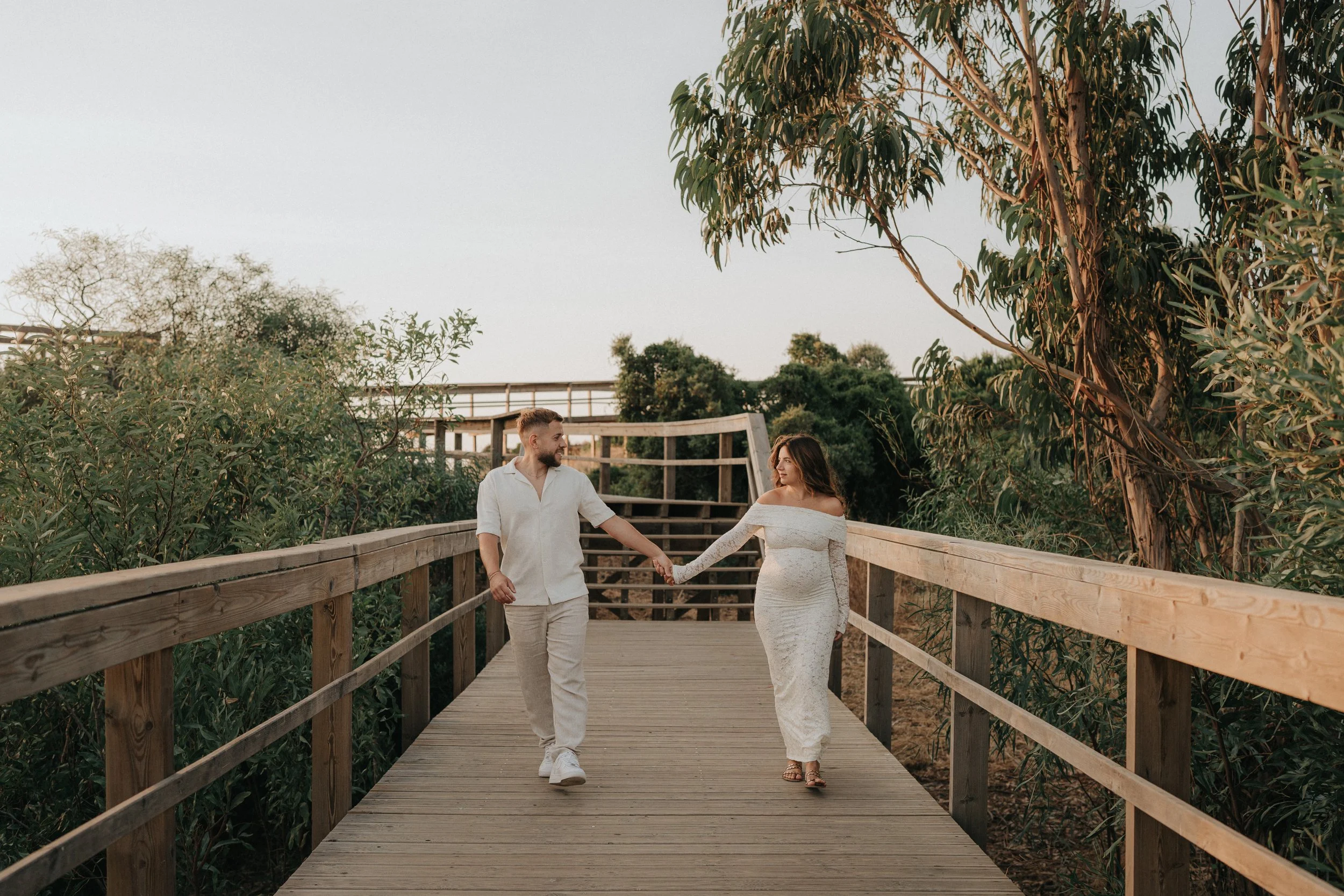 A couple is walking hand in hand on a wooden bridge surrounded by lush greenery, with the man in a white shirt and the woman in a white dress.