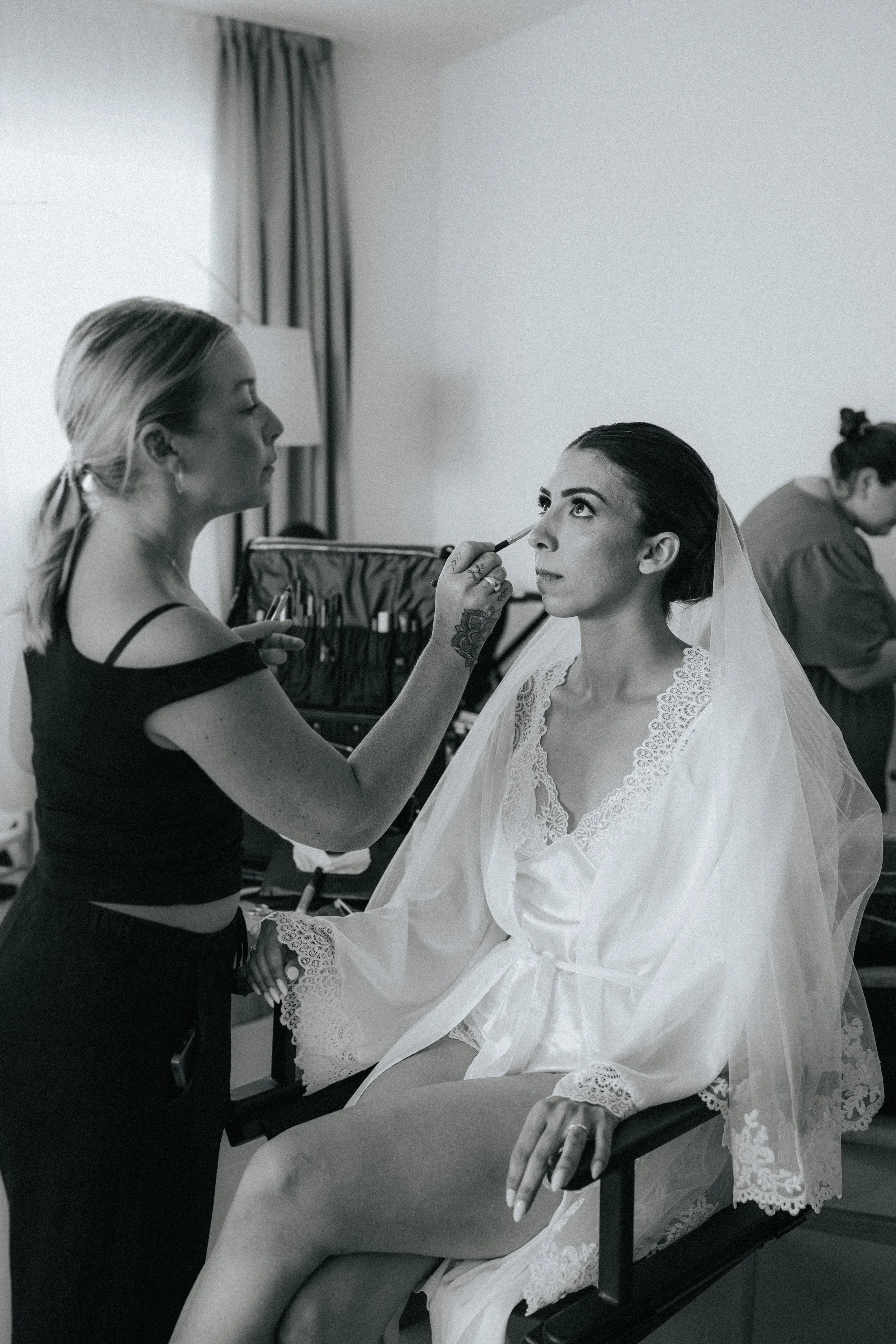 A makeup artist applies makeup to a bride sitting on a bed, with another person in the background.