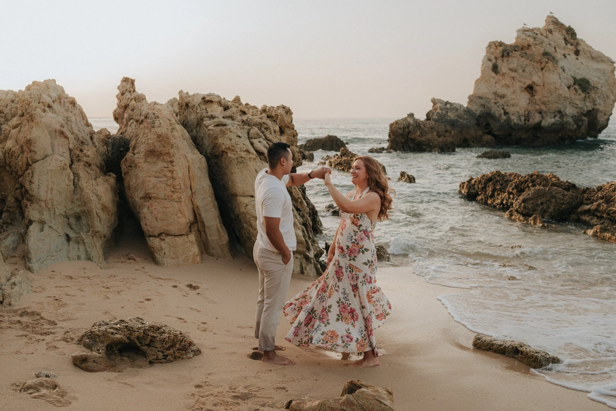 A couple holding hands on a sandy beach near rocky formations, with the ocean in the background and a woman in a floral dress smiling at her partner.