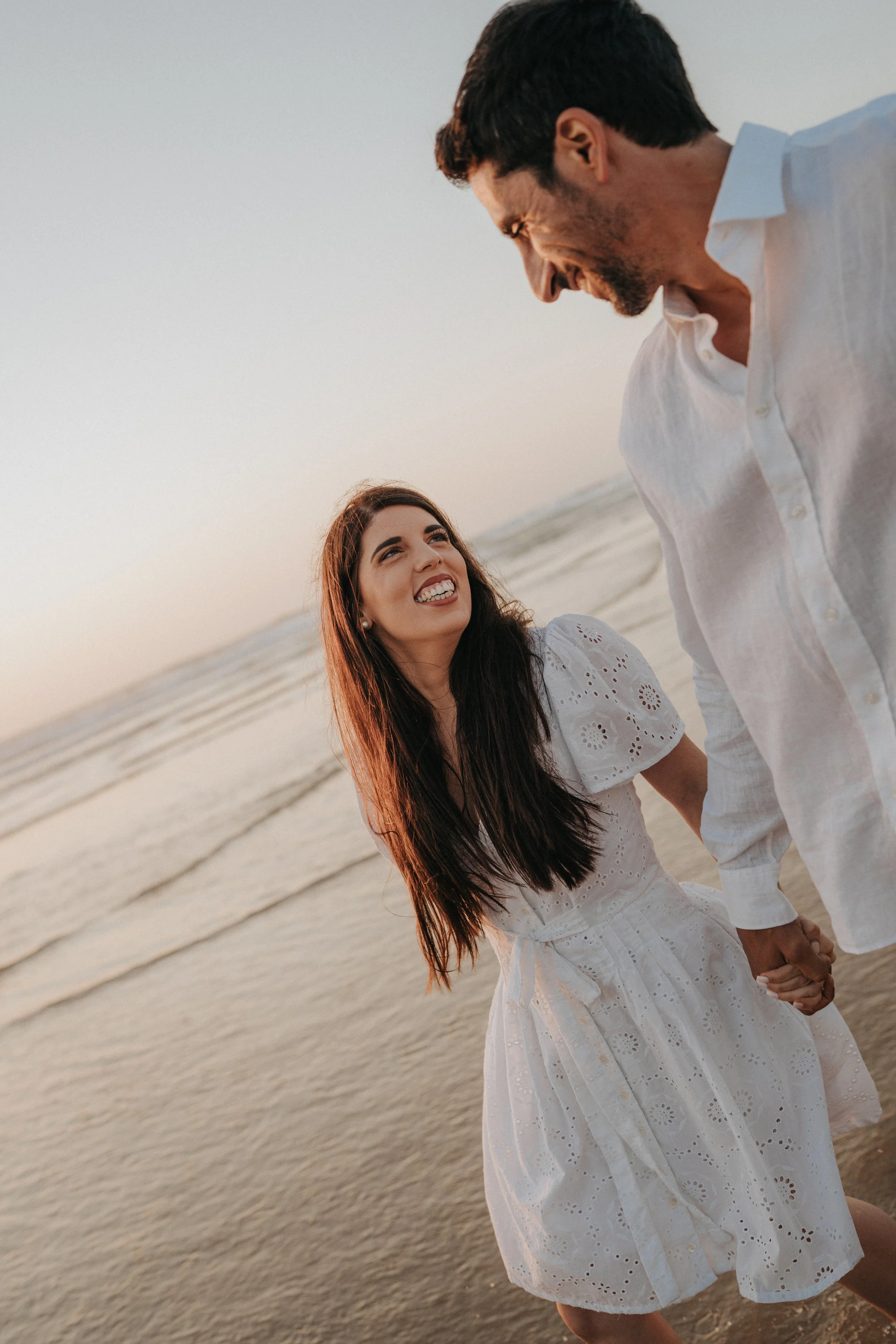 A smiling woman with long dark hair wearing a white dress and a smiling man in a white shirt standing at the beach during sunset, holding hands.