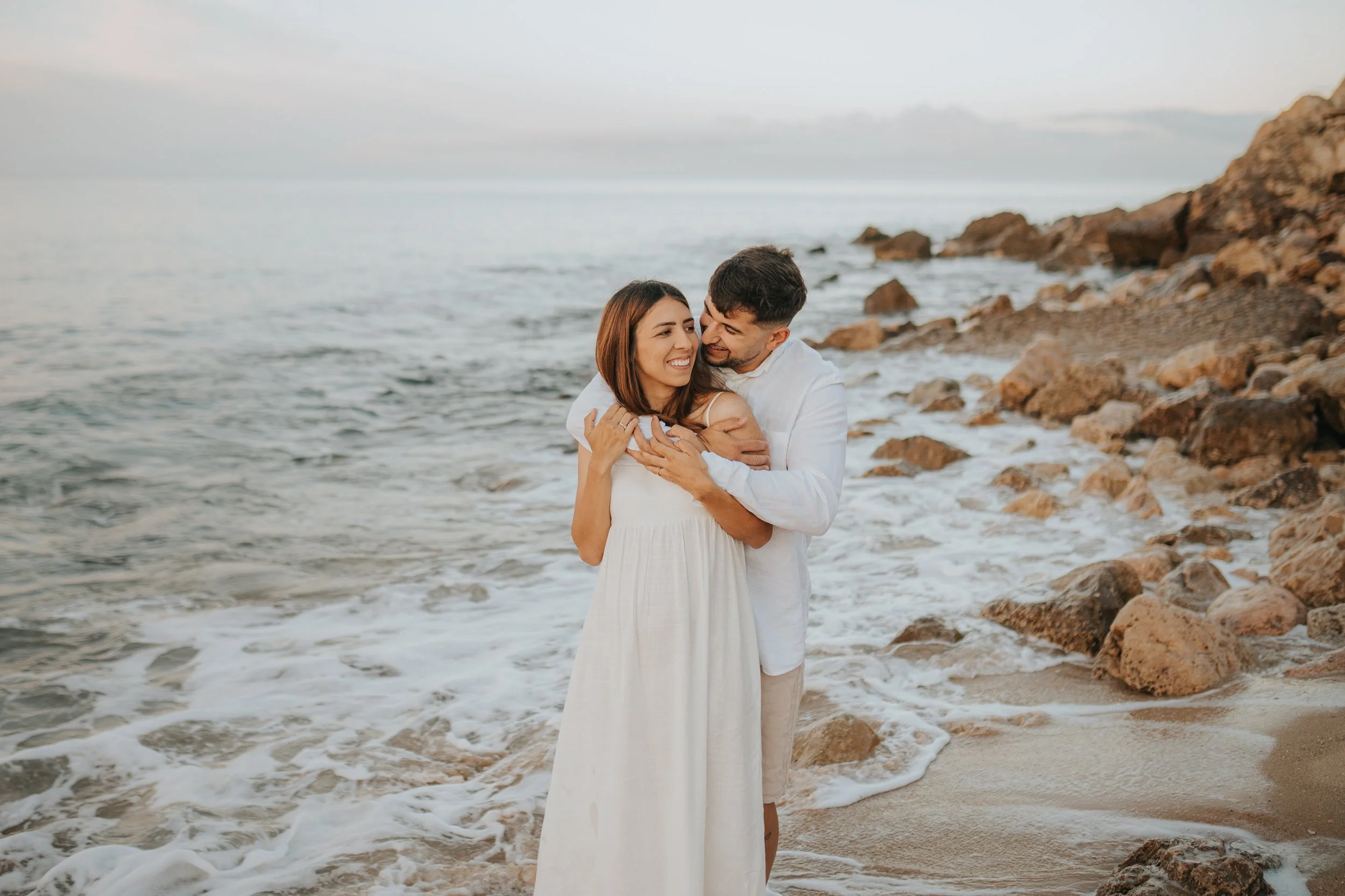 A couple embracing and smiling on a rocky beach, with waves and a cloudy sky in the background.