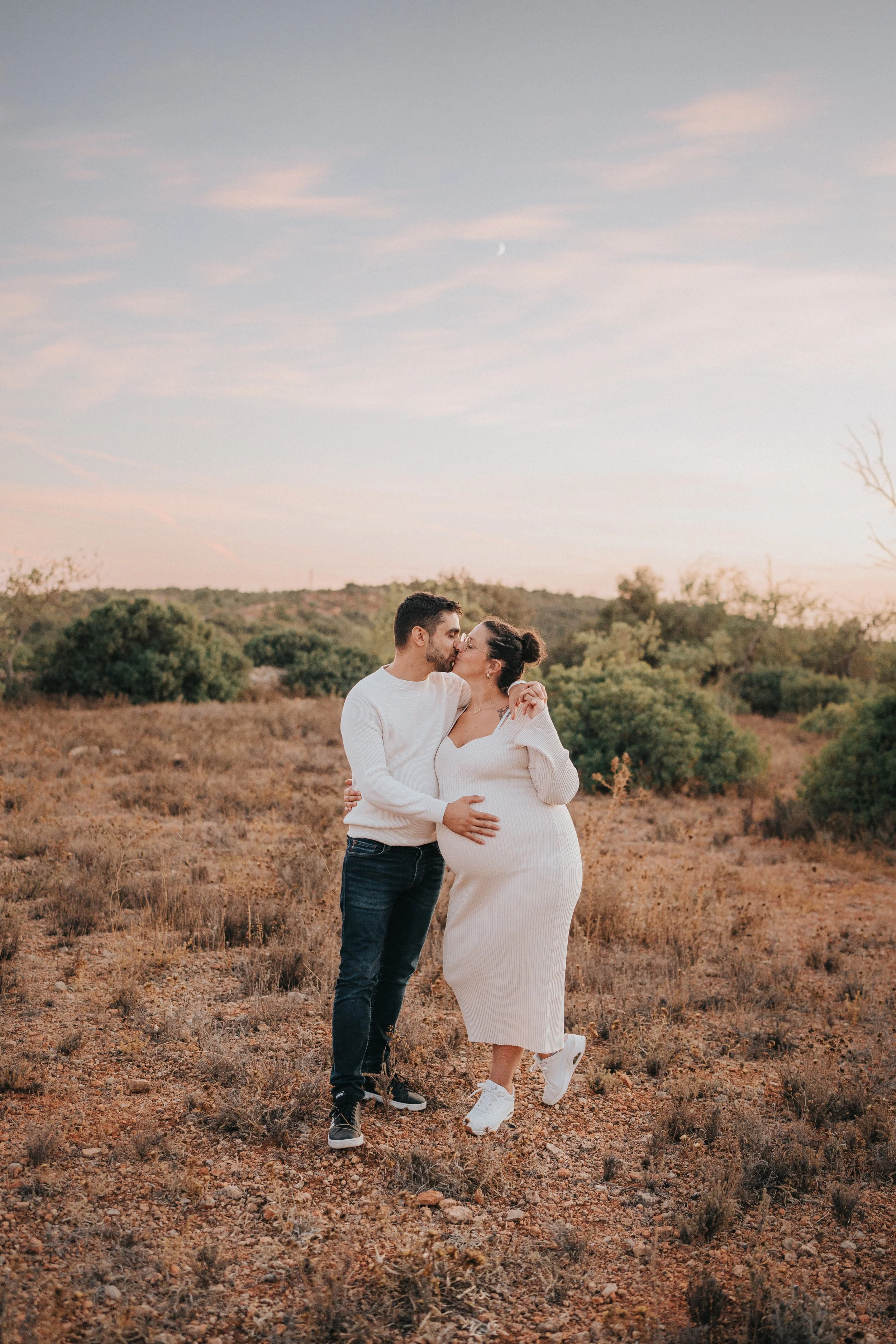 A couple stands in a desert landscape at sunset, embracing and sharing a kiss, with the woman pregnant and wearing a white dress and sneakers and the man in a white sweater, jeans, and sneakers.