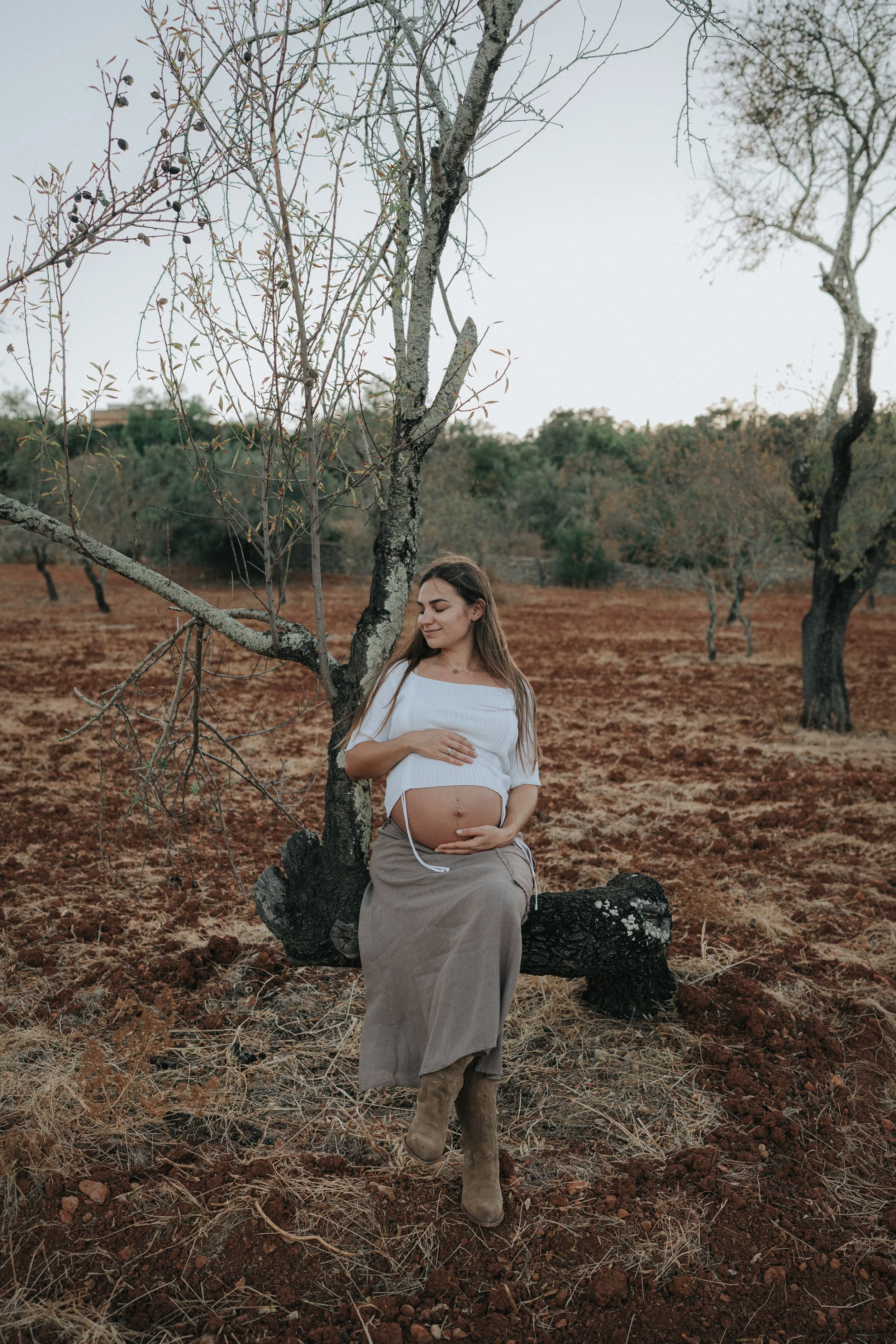 A pregnant woman sitting on a fallen tree trunk outdoors, holding her belly with both hands, surrounded by bare trees and a dry, brown landscape.